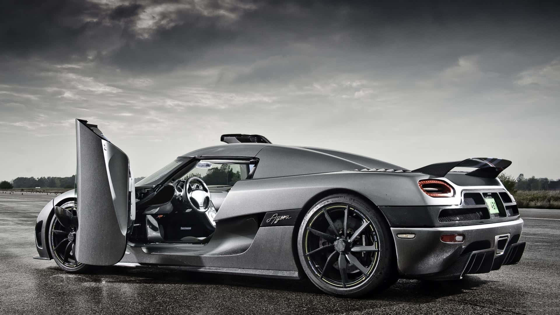 高清 桌布 壁紙 — 一架流線型銀色Koenigsegg Agera，車門向上打開，停在濕滑的瀝青賽道上，背景是戲劇性的多雲天空。