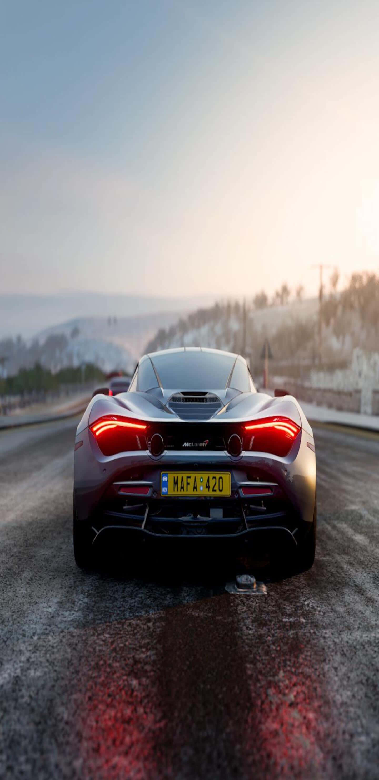 Pixel 3xl Mclaren 720s 桌布 壁紙 — 一架流線型銀色McLaren跑車，紅色尾燈閃爍，在日落時分行駛在濕滑的道路上，穿越白雪覆蓋的山區景色。