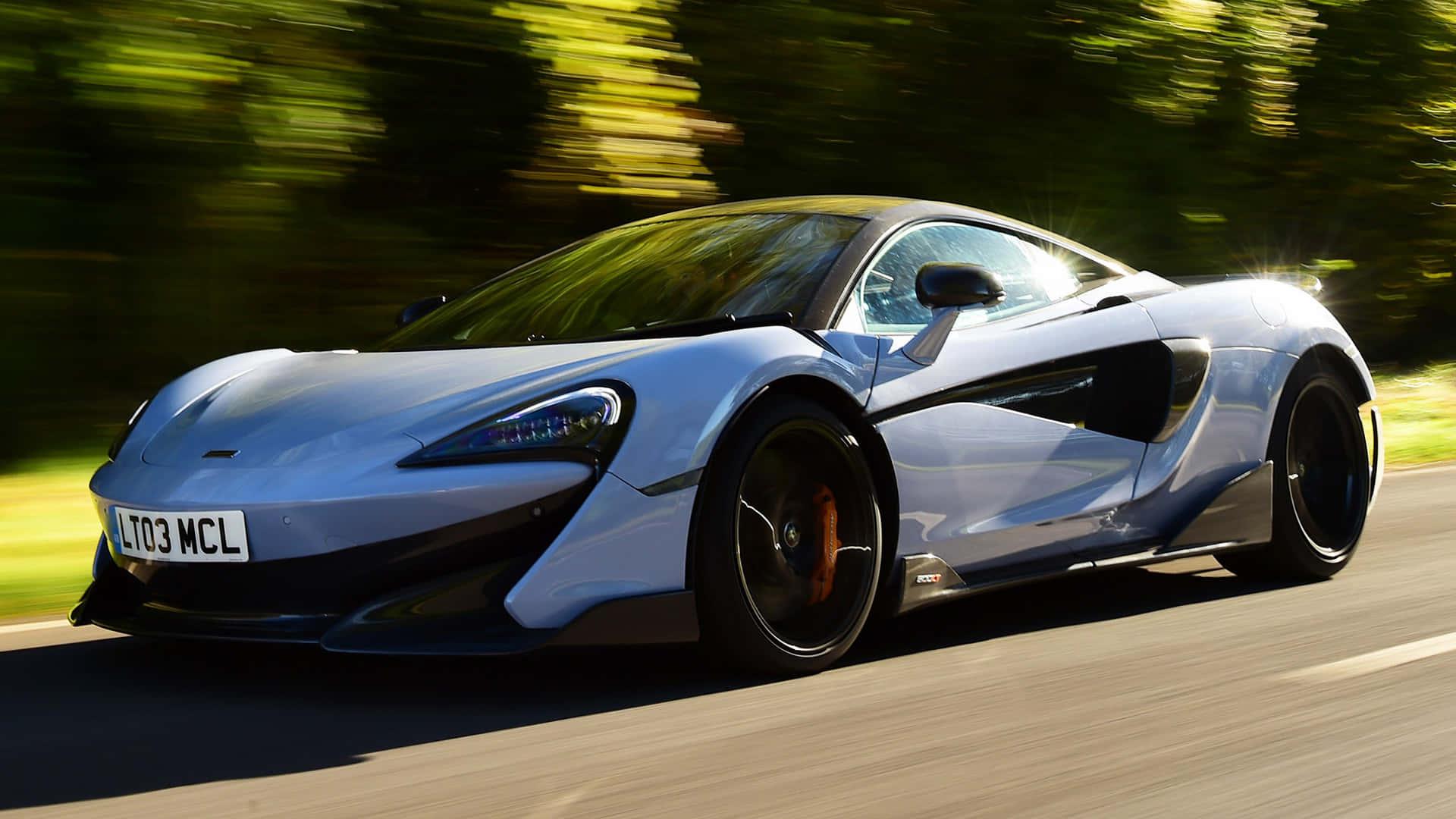 麥拉倫 桌布 壁紙 — 一架流線型銀色McLaren跑車在陽光明媚的道路上行駛，背景是模糊的綠色樹葉。