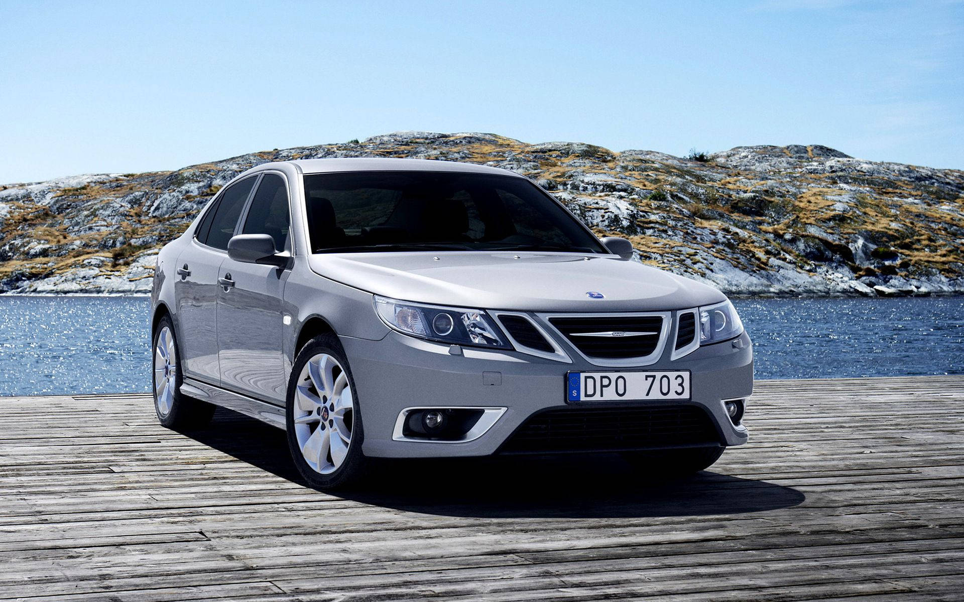 Saab 桌布 壁紙 — 一架流線型銀色Saab轎車停泊在風化的木碼頭上，旁邊是平靜的藍色海水，背景是岩石丘陵，天空晴朗。