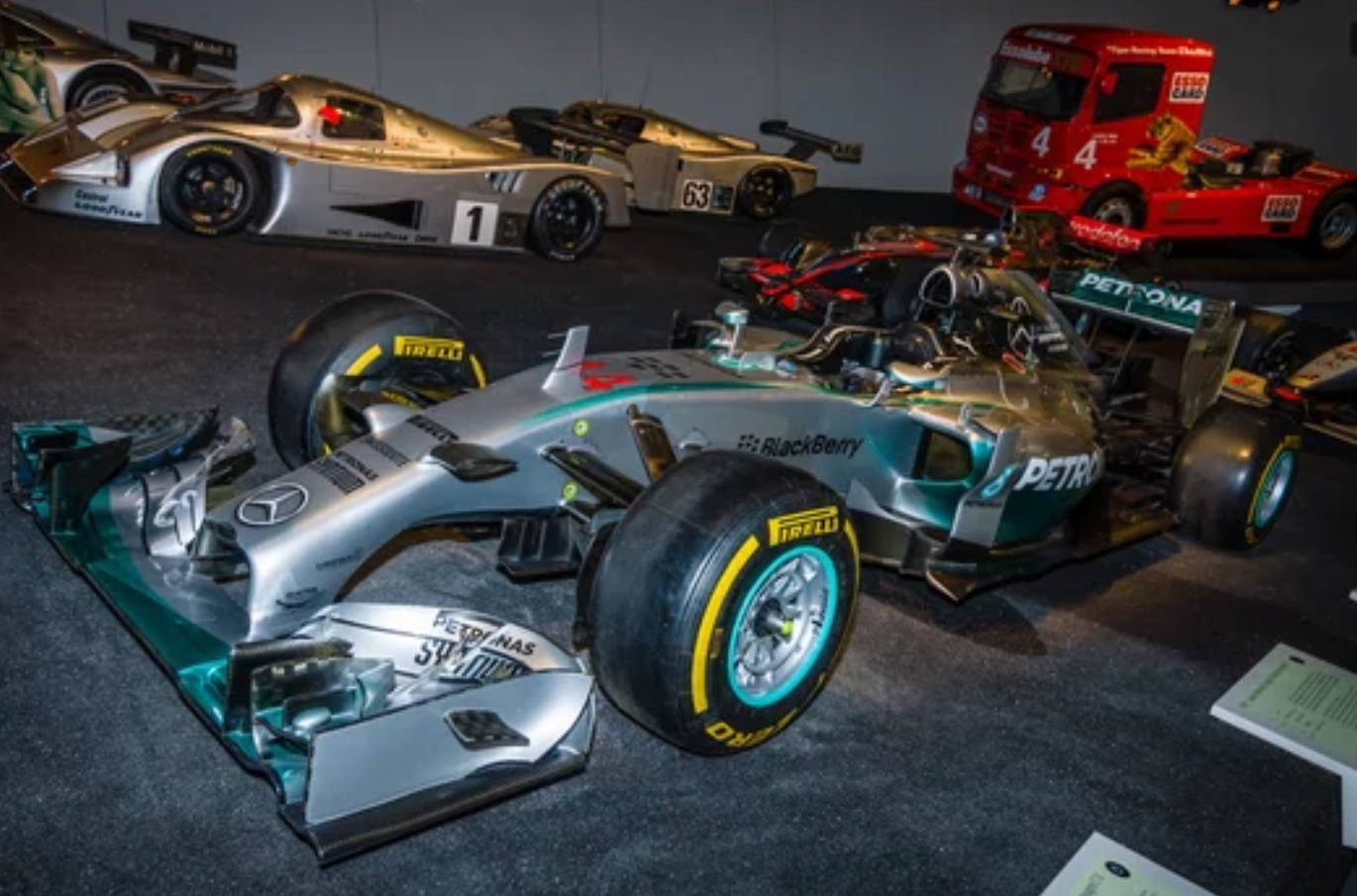 Mercedes 桌布 壁紙 — 一架流線型銀色和青綠色的 Formula 1 賽車配備 Pirelli 輪胎，在博物館內與其他賽車一同於人造燈光下展出。