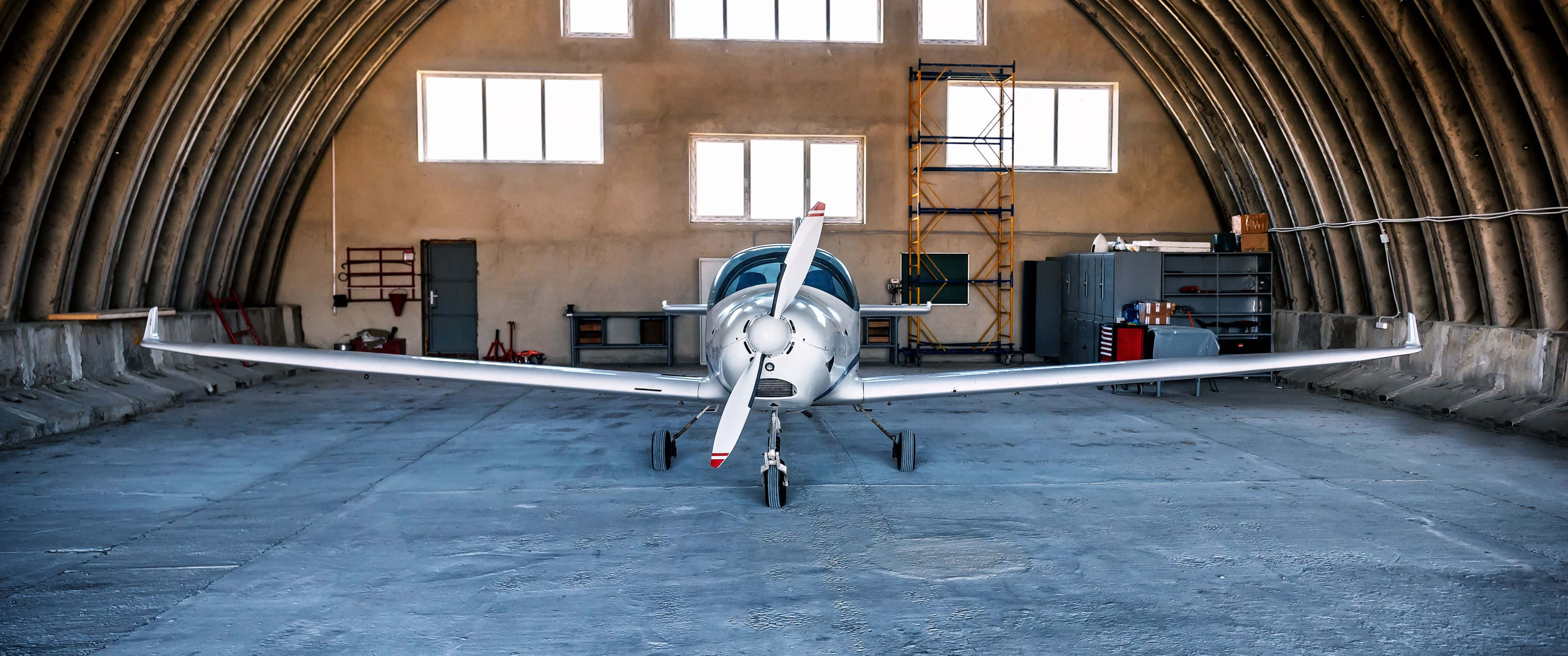 3440x1440p Small Planes 桌布 壁紙 — 一架流線型銀色單引擎飛機停放在寬敞的拱形機庫中央，機庫有混凝土地板和高窗。