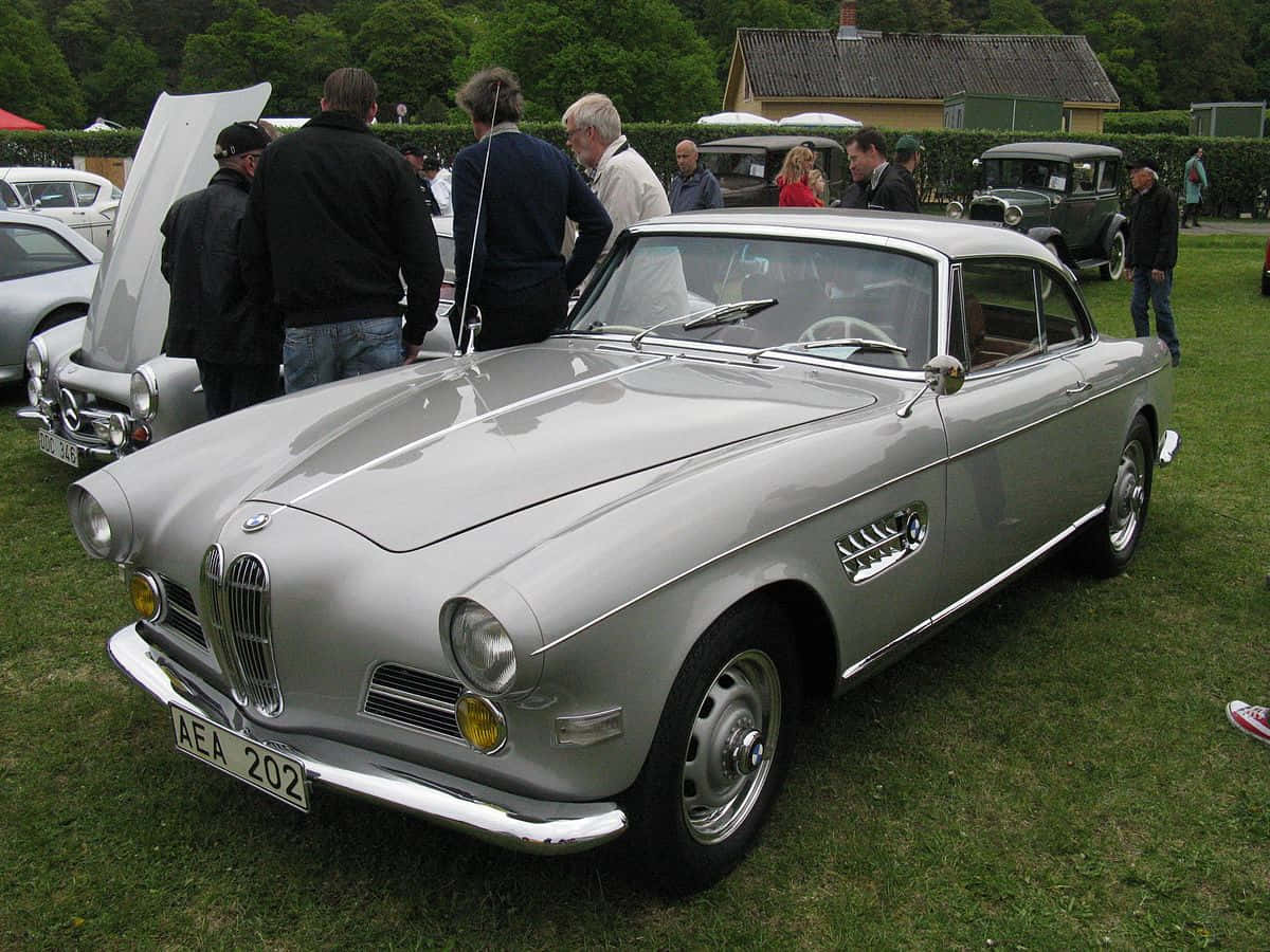 BMW 動圖 桌布 壁紙 — 一架流線型銀色復古BMW 507跑車在戶外經典車展的日光下閃耀，周圍有觀眾聚集。