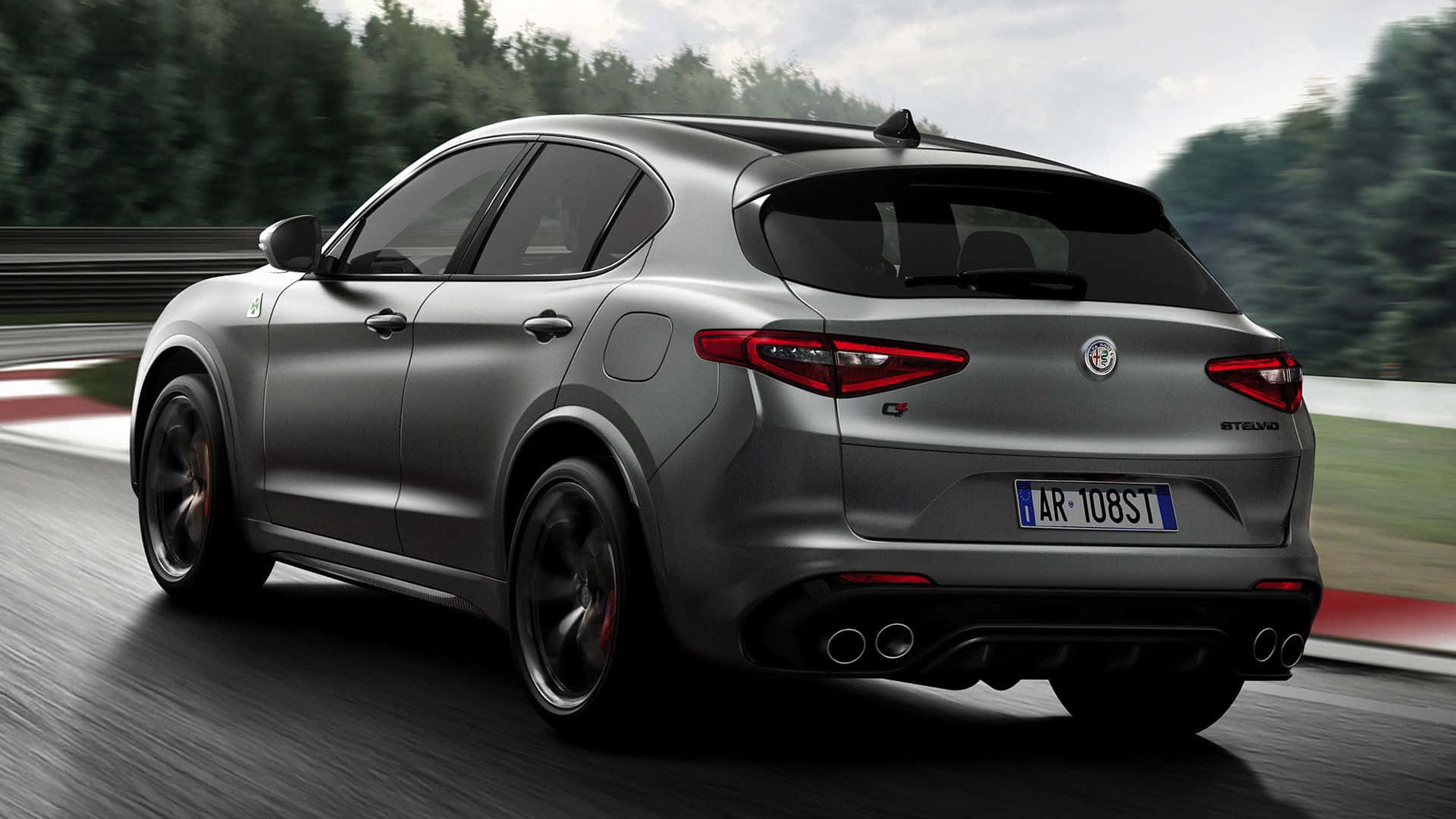 Alfa Romeo Stelvio 桌布 壁紙 — 一架流線型銀色愛快羅密歐 Stelvio Quadrifoglio 在賽道上飛馳，背景是模糊的綠色樹木和紅白路緣。