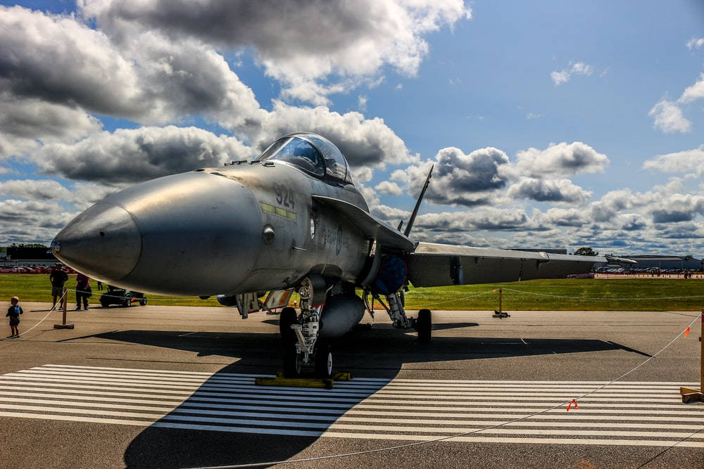 Military Jets 桌布 壁紙 — 一架流線型銀色戰鬥機停在機場停機坪上，天空多雲而富有戲劇性，附近有人群。