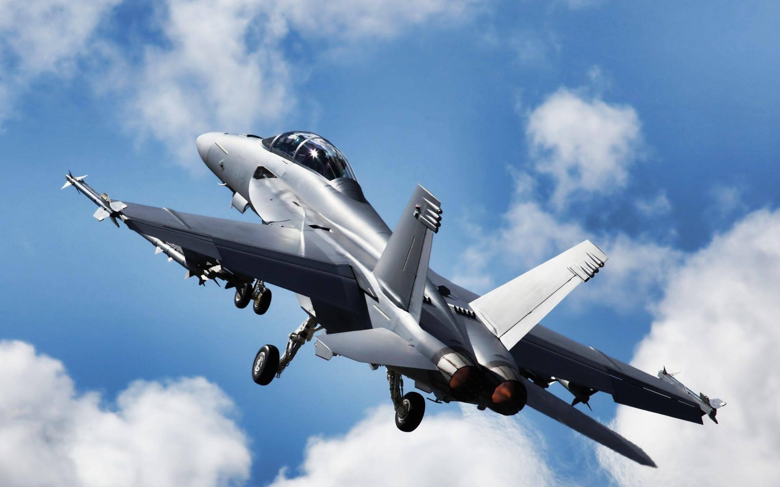 軍事動圖 桌布 壁紙 — 一架流線型銀色戰鬥機在點綴著蓬鬆白雲的明亮藍天中翱翔。
