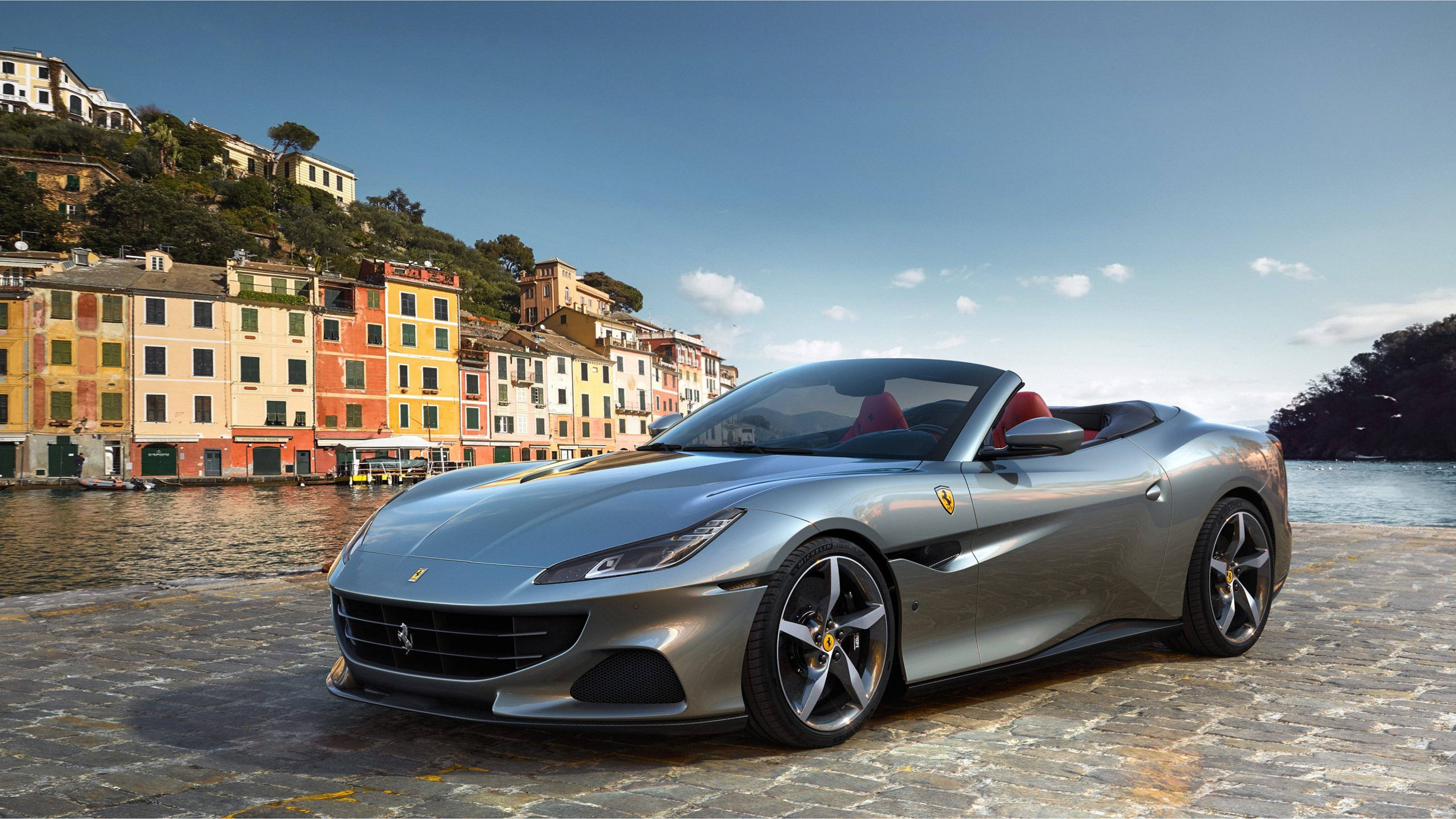 跑車 桌布 壁紙 — 一架流線型銀色法拉利 Portofino 敞篷車停泊在鵝卵石海濱，背景是色彩繽紛的義大利山丘建築和寧靜的水面。