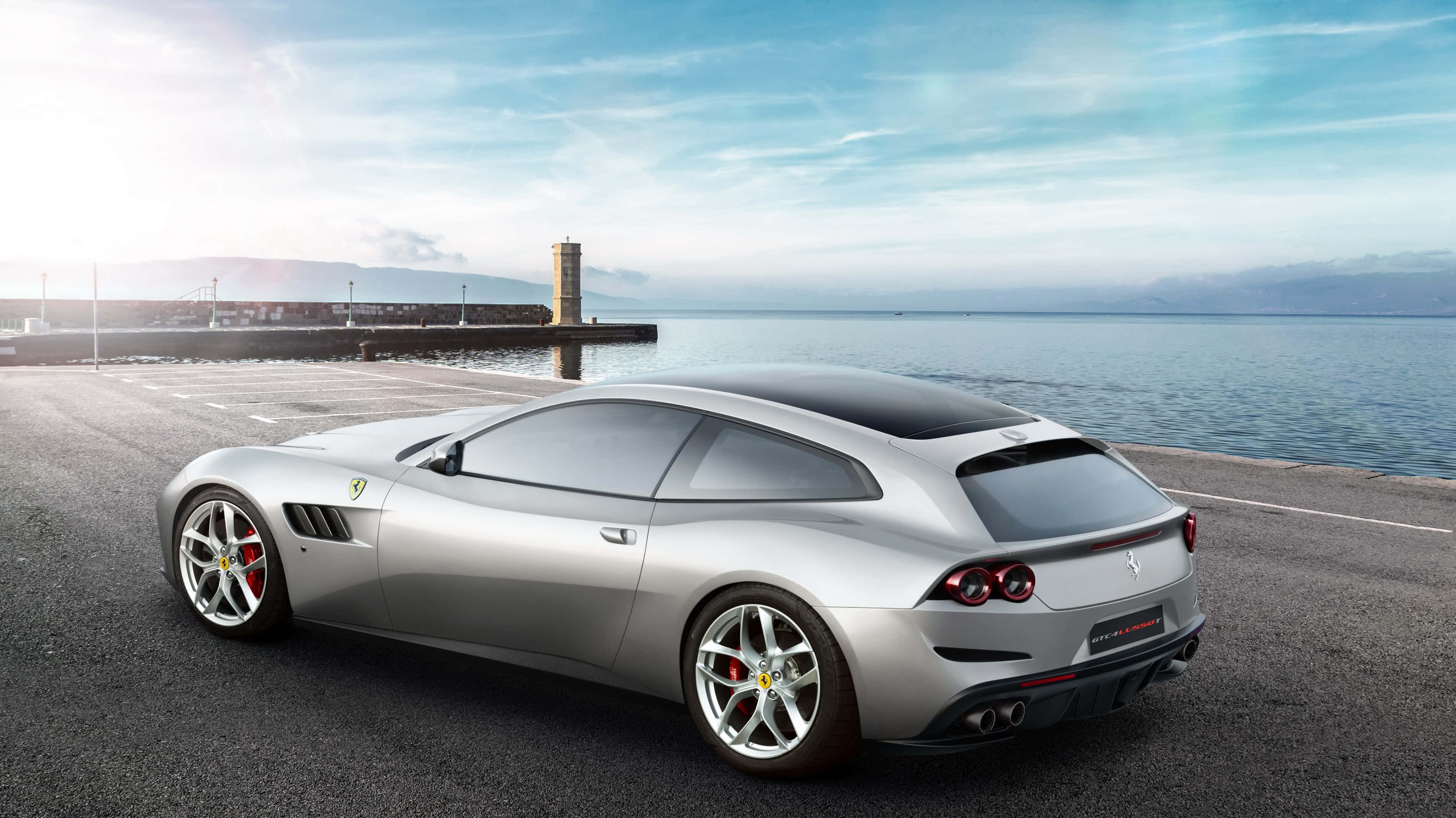 Ferrari Gtc4lusso 桌布 壁紙 — 一架流線型銀色法拉利跑車停泊在海岸碼頭上，背景是平靜的海面、遠山和明亮的藍天。