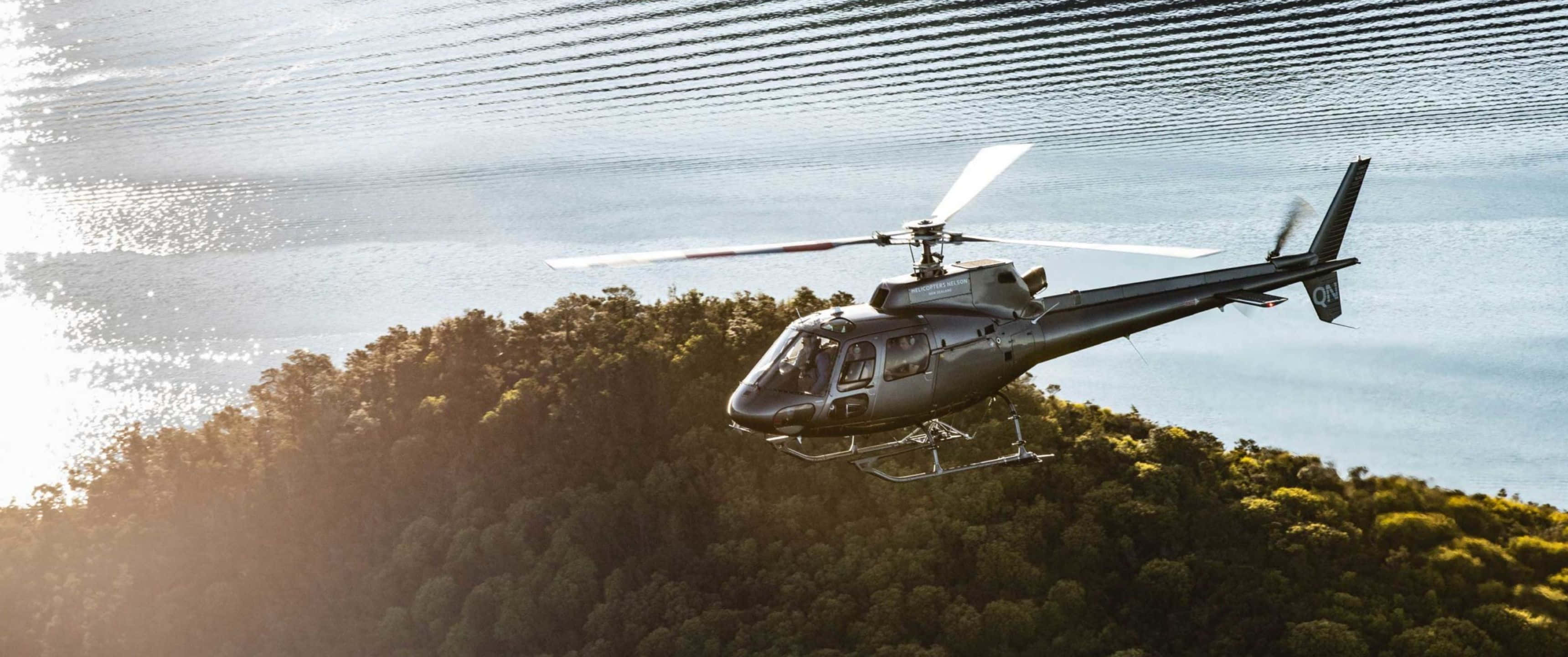 3440x1440p Helicopters 桌布 壁紙 — 一架流線型銀色直升機低飛過陽光明媚的樹林山丘，旁邊是波光粼粼的湖泊，天空帶有條紋。