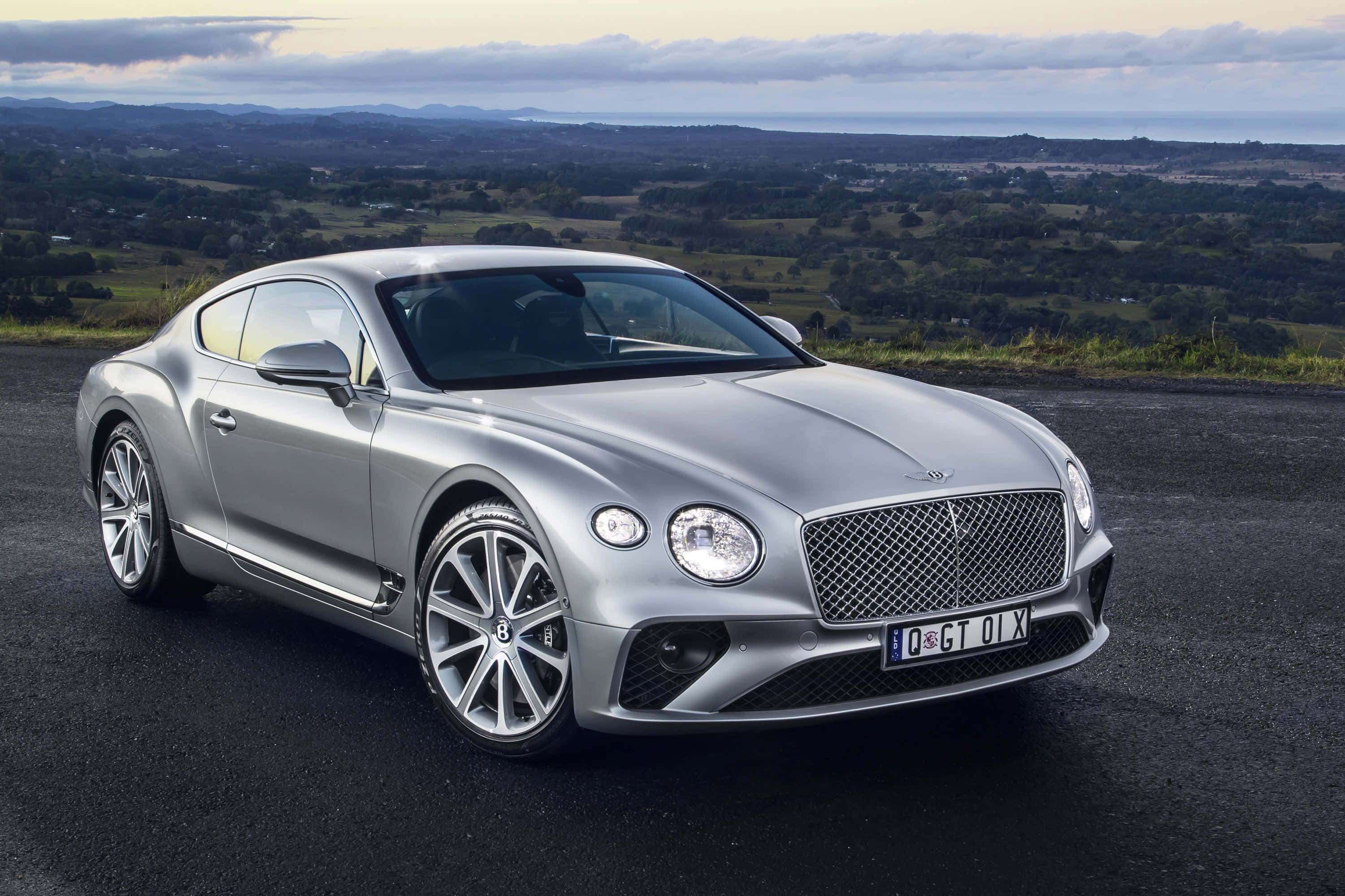 賓利 桌布 壁紙 — 一架流線型銀色賓利 Continental GT 轎跑車停泊在昏暗的道路上，俯瞰廣闊的綠色山谷，天空多雲。