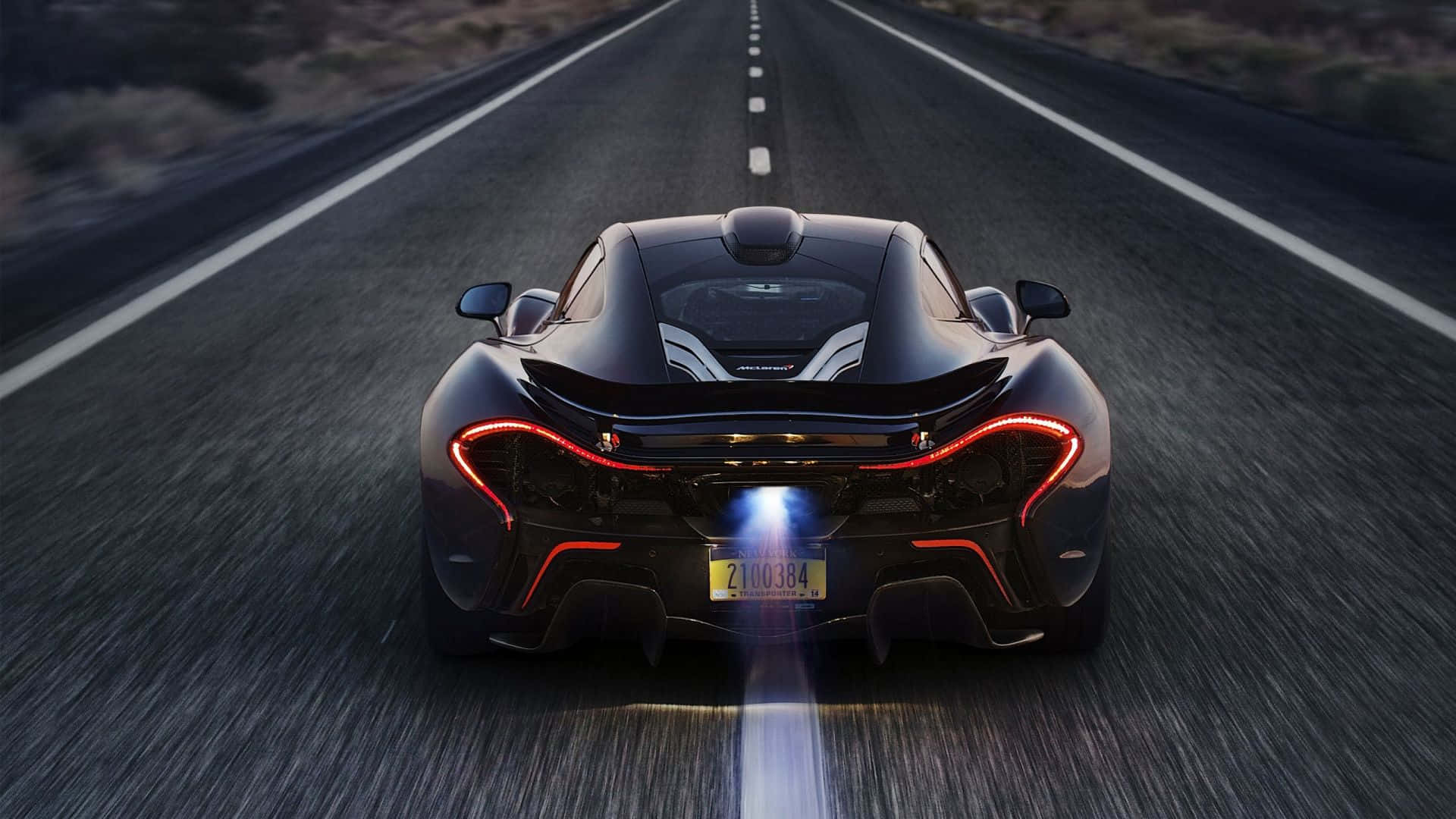 桌布 壁紙 — 一架流線型黑色McLaren P1在黃昏時分於沙漠公路疾馳，帶有發光的紅色尾燈和明亮的排氣火焰。