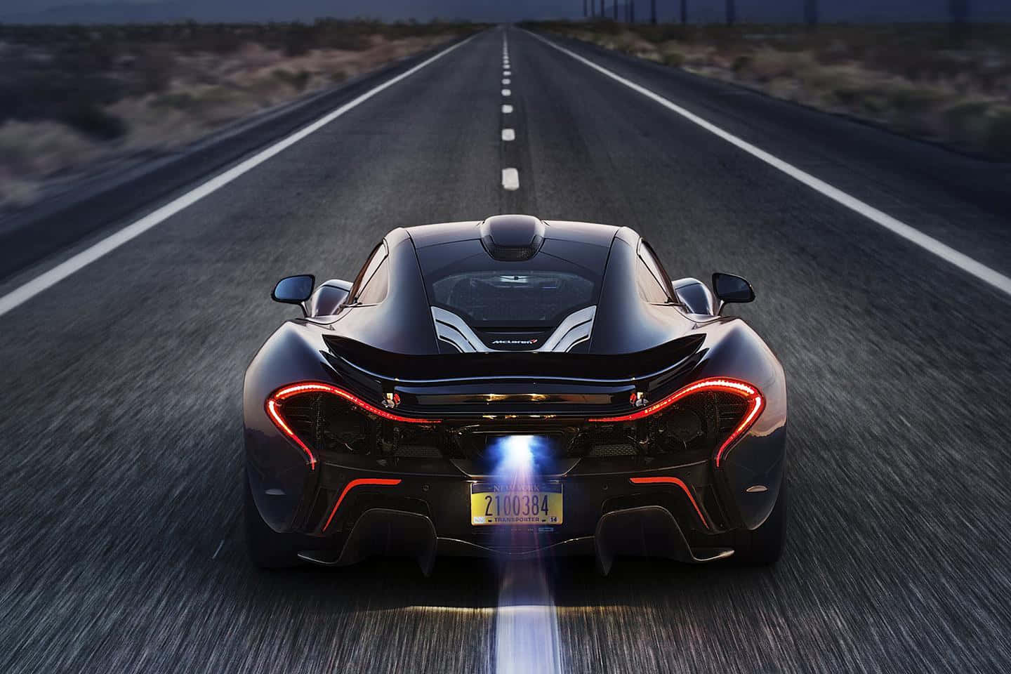 麥拉倫 桌布 壁紙 — 一架流線型黑色McLaren P1在黃昏時分沿著筆直的沙漠高速公路飛馳，帶有發光的紅色尾燈和藍色排氣火焰。