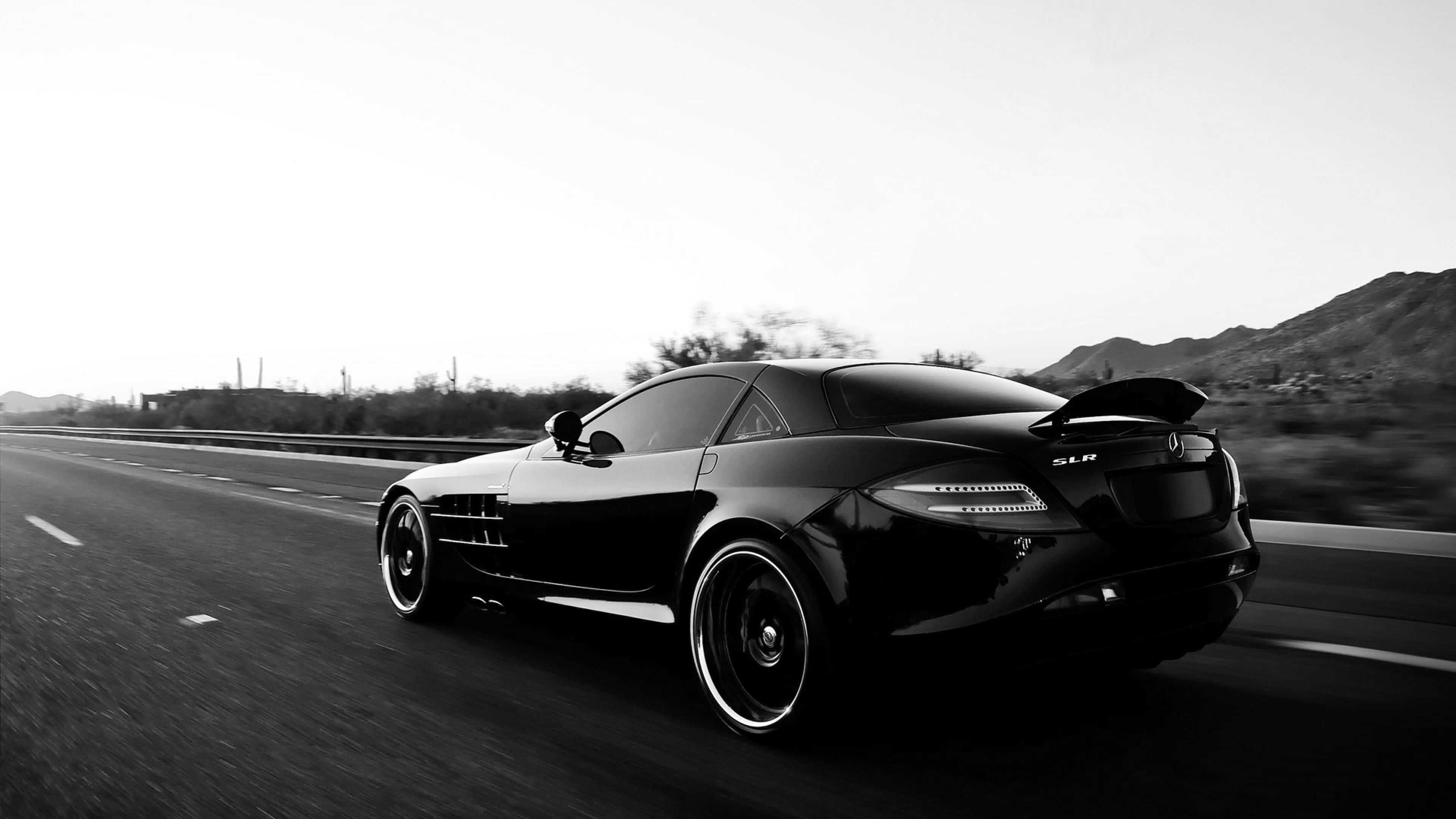 Black Car 4K 桌布 壁紙 — 一架流線型黑色梅賽德斯 SLR 麥拉倫在朦朧天空下，以模糊動態和遠山為背景，在沙漠高速公路上飛馳。