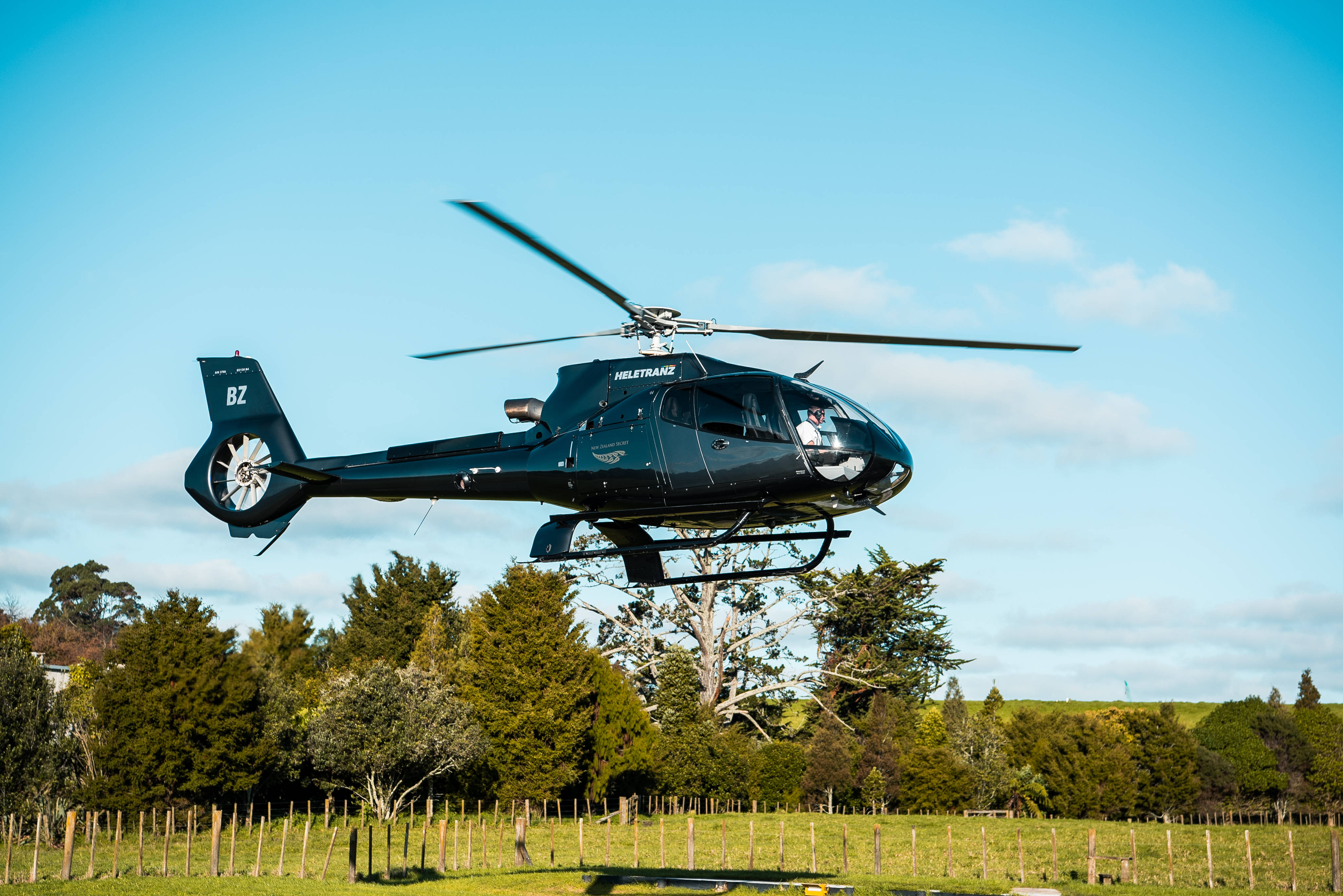 Helicopter 桌布 壁紙 — 一架流線型黑色直升機在陽光明媚的綠色田野上空盤旋，田野上有樹木和圍欄，背景是清澈的藍天。