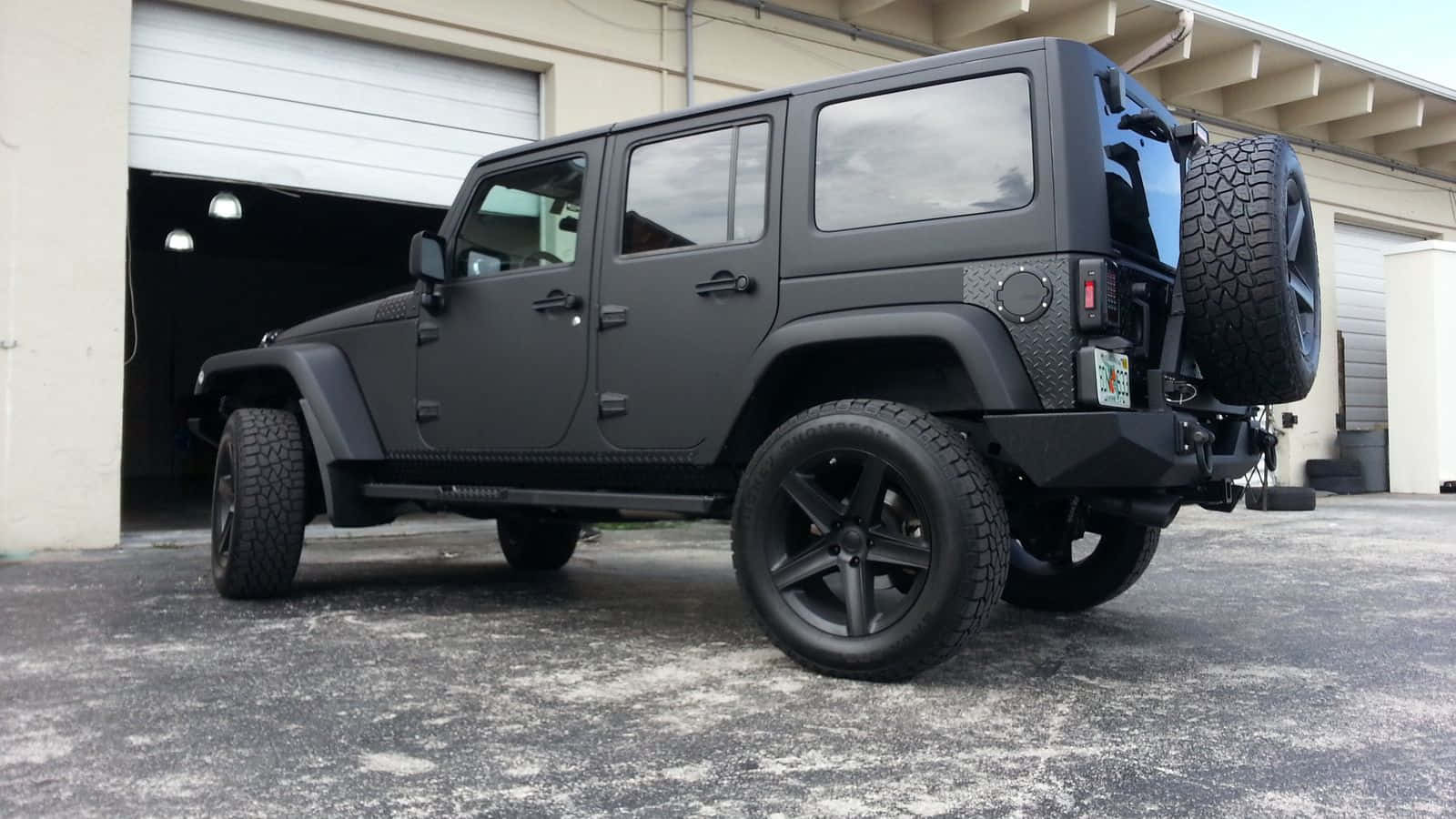 黑色吉普車 桌布 壁紙 — 一架消光黑色Jeep Wrangler配備超大輪胎，停在混凝土場地上的車庫前。