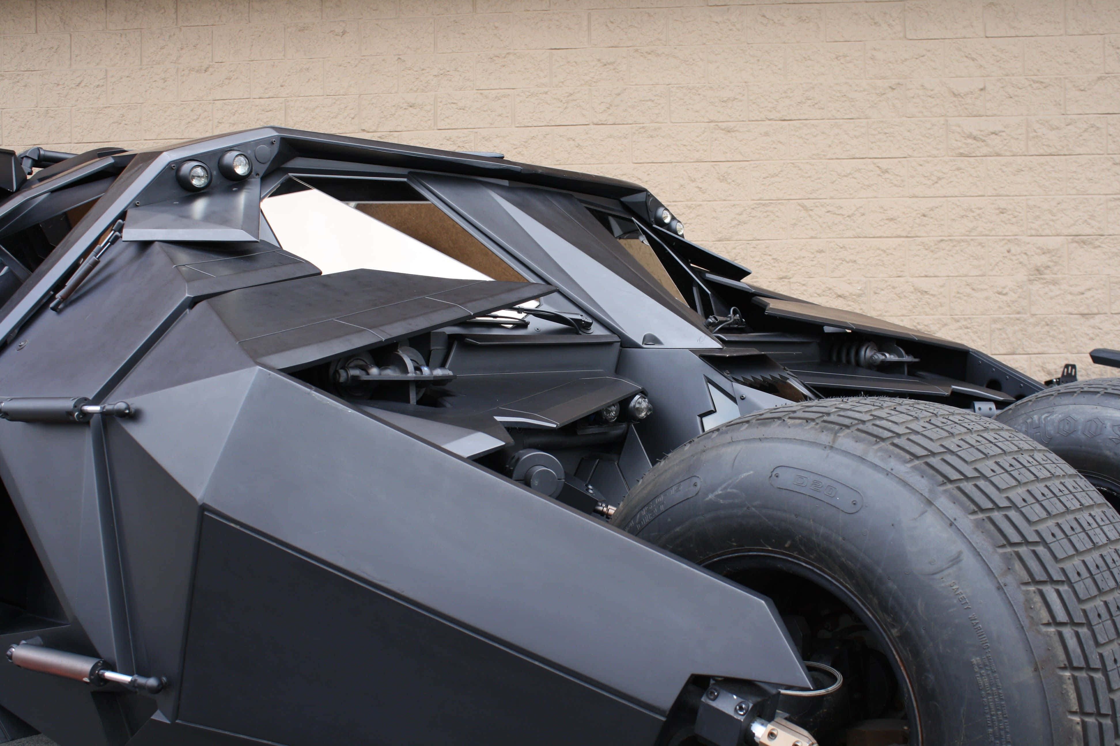 4K Batmobile 桌布 壁紙 — 一架消光黑色裝甲蝙蝠車，採用棱角設計和大輪胎，停泊在米色牆壁前的特寫。