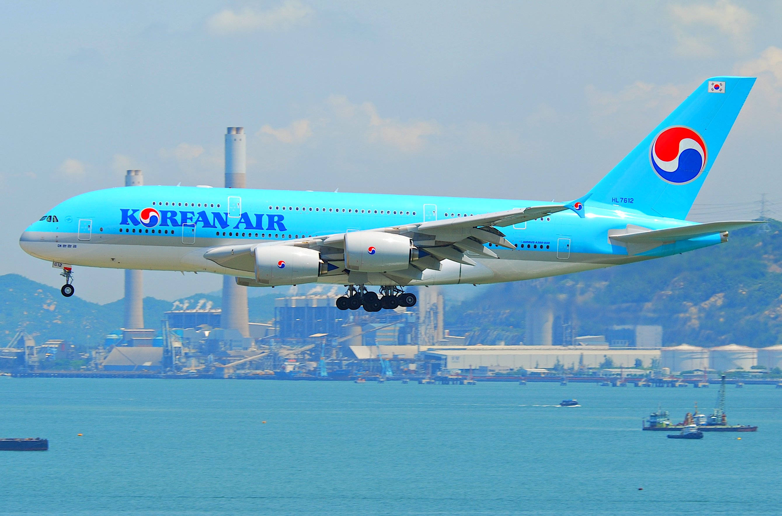 風景 桌布 壁紙 — 一架淺藍色塗裝的大韓航空空中巴士A380在工業海岸線和山丘背景下，優雅地降落在碧綠的水面上。
