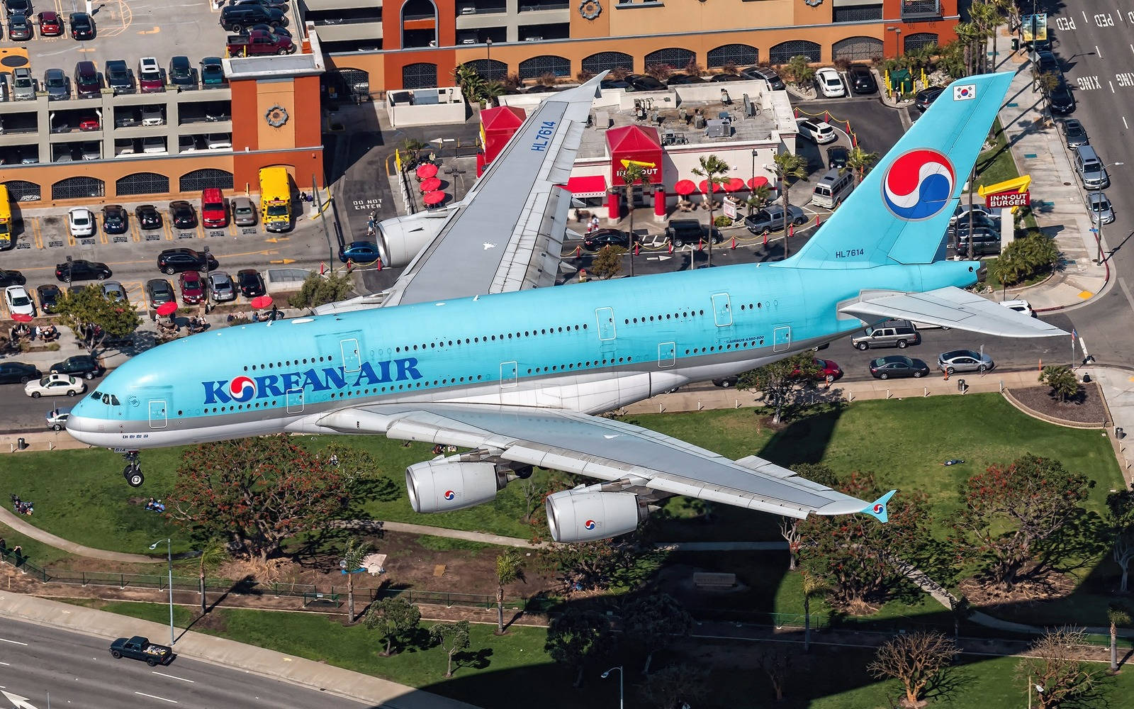 飛機 桌布 壁紙 — 一架淺藍色塗裝的大韓航空空中巴士A380在陽光普照的城市景觀上空低飛，下方有停車場和建築物。