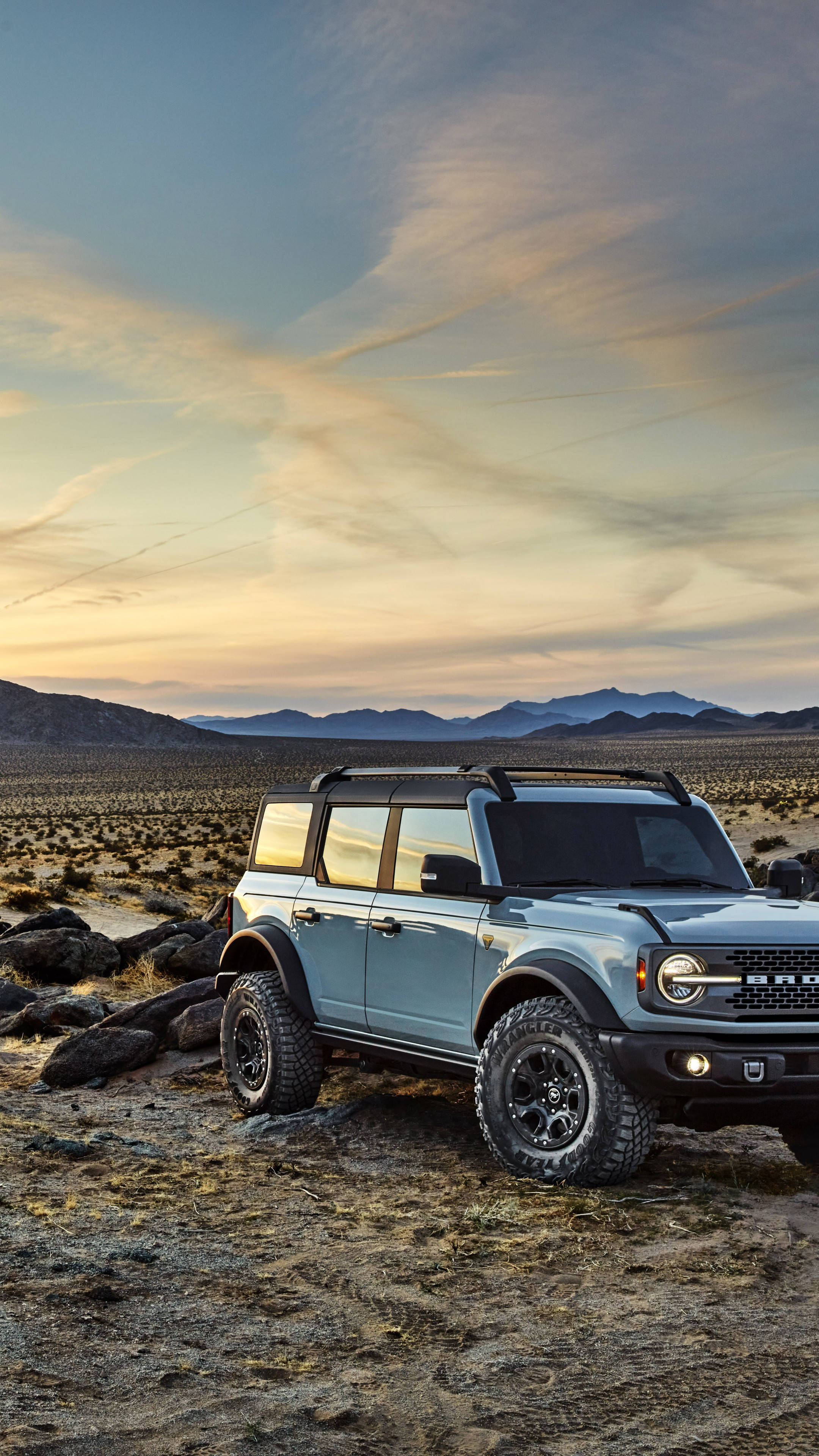 Ford Bronco 桌布 壁紙 — 一架淺藍色福特Bronco在日落時分堅固地矗立在岩石沙漠地形上，遠山在條紋天空下。