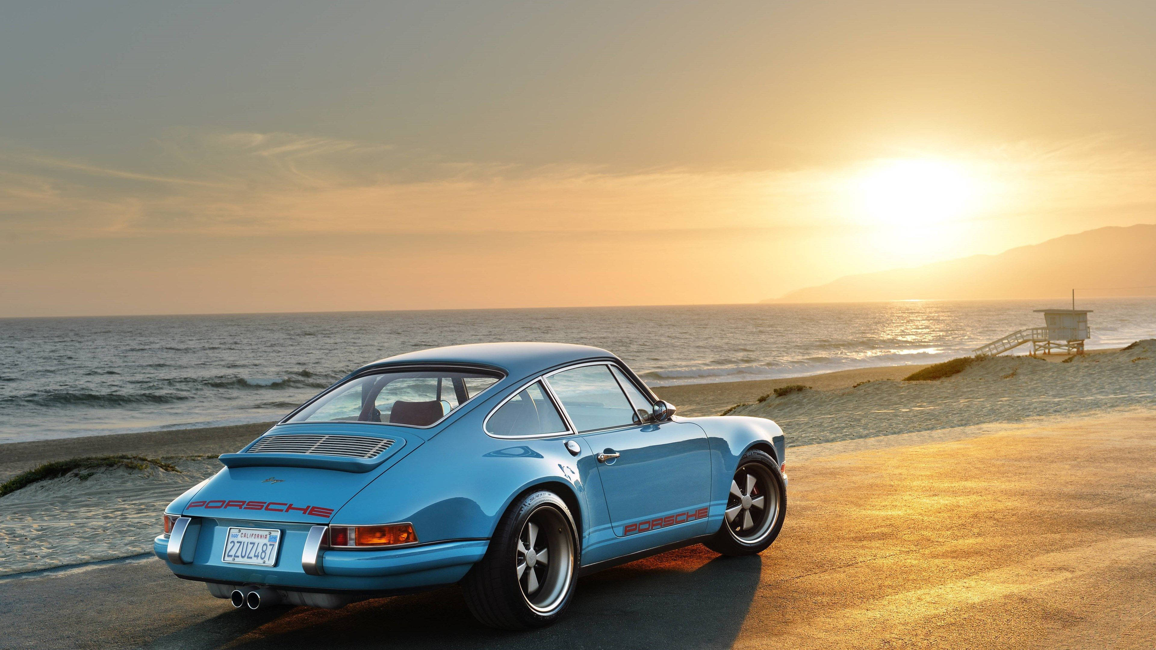 Singer 桌布 壁紙 — 一架淺藍色經典保時捷911停泊在日落時分的海岸公路上，背景有海浪和救生塔。