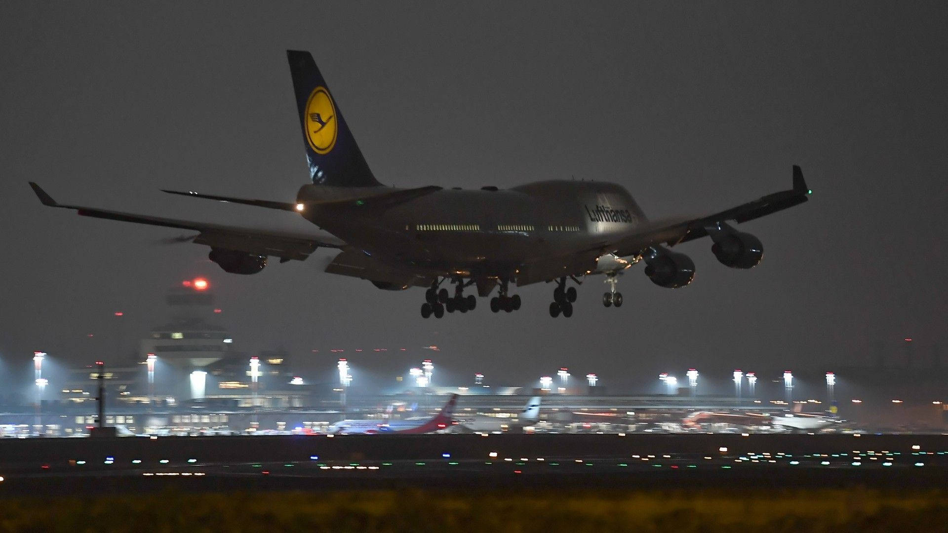夜空 桌布 壁紙 — 一架漢莎航空波音747在夜間放下起落架，以燈火通明的機場航廈為背景下降。