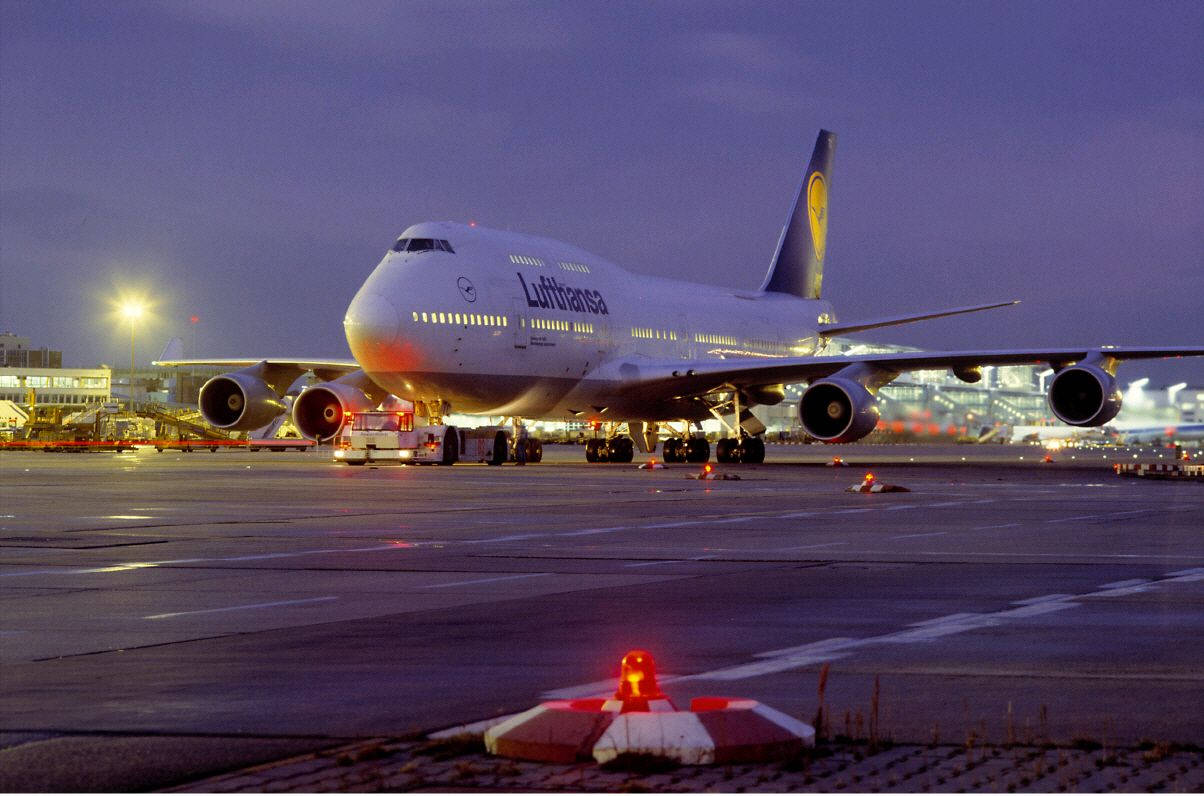 飛機 桌布 壁紙 — 一架漢莎航空波音747在暮色燈光下於機場停機坪發光，前景有紅色信標。