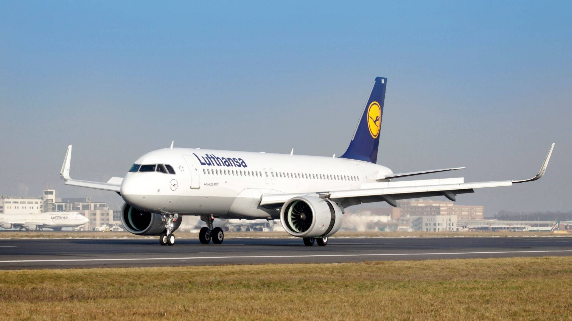 飛機 桌布 壁紙 — 一架漢莎航空空中巴士A320在晴朗藍天下滑行，遠處朦朧可見機場建築。
