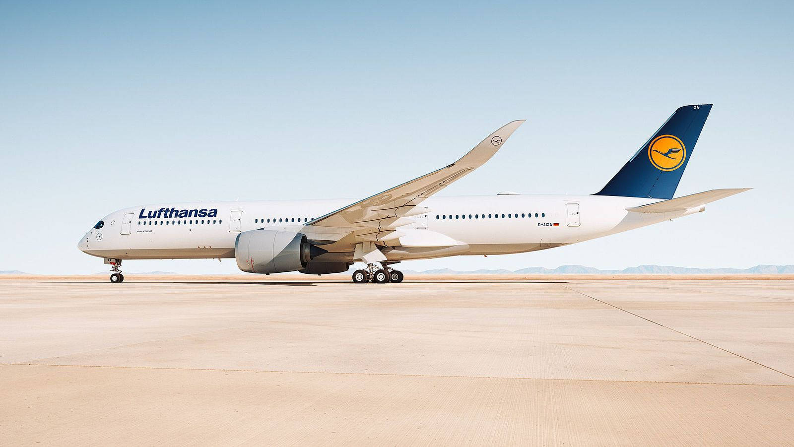  壁紙 — 一架漢莎航空空中巴士A350停泊在陽光普照的停機坪上，背景是清澈的藍天和遠山。