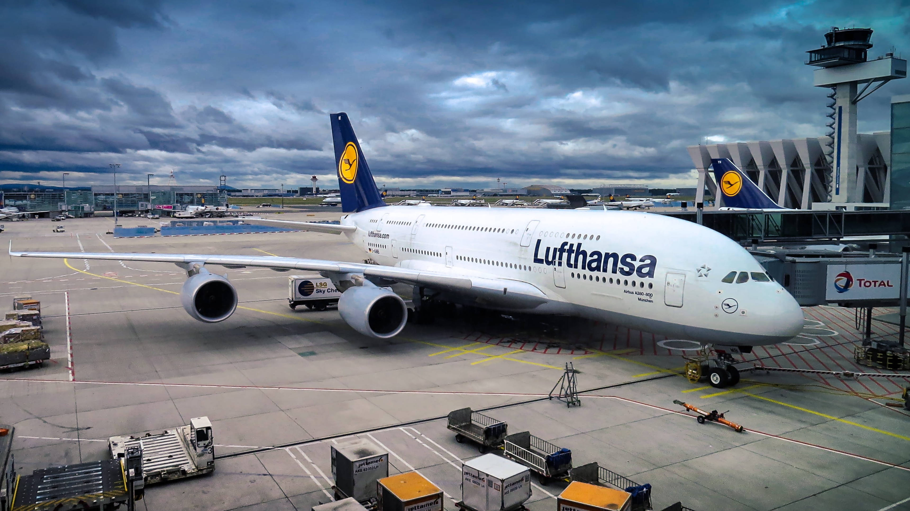 Airport 桌布 壁紙 — 一架漢莎航空空中巴士A380停泊在機場閘口，天空多雲，氣氛戲劇化，附近有地面服務設備。