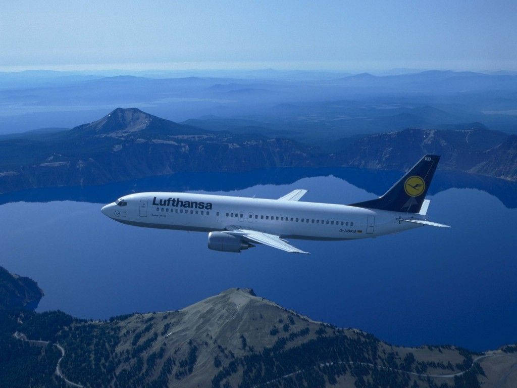 山脈 桌布 壁紙 — 一架漢莎航空飛機在晴朗的天空下，優雅地翱翔於崎嶇山脈環繞的廣闊藍色湖泊之上。