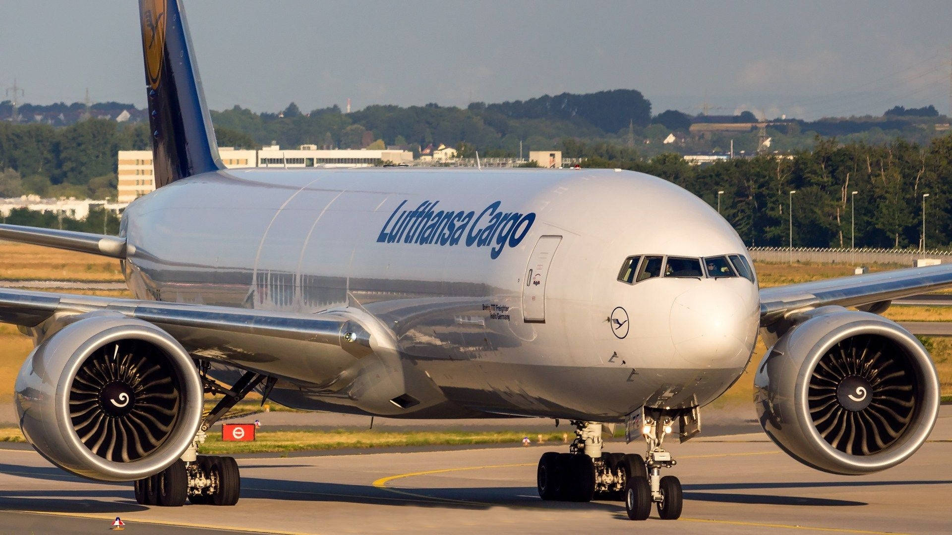 飛機 桌布 壁紙 — 一架漢莎貨運波音777停放在機場停機坪上，背景有樹木和建築物，天空晴朗。
