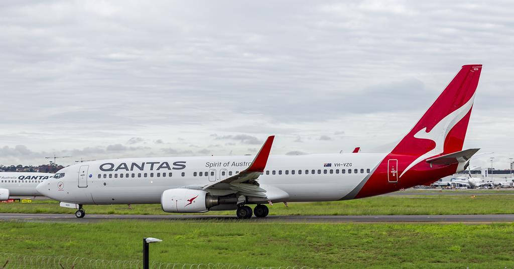  壁紙 — 一架澳洲航空Qantas Spirit of Australia的波音737在潮濕的跑道上滑行，天空多雲，背景有綠色草地和機場基礎設施。