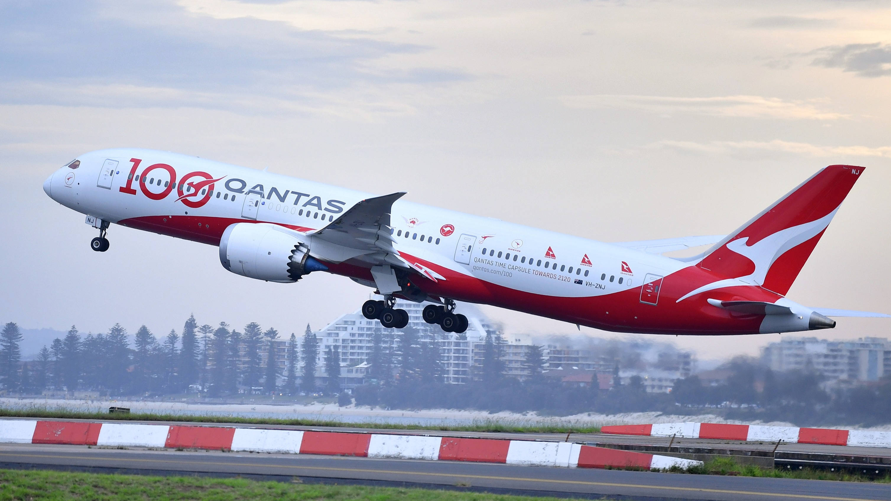 飛機 桌布 壁紙 — 一架澳洲航空波音787在黃昏時分放下起落架，在陰天和遠處的城市景觀中起飛。