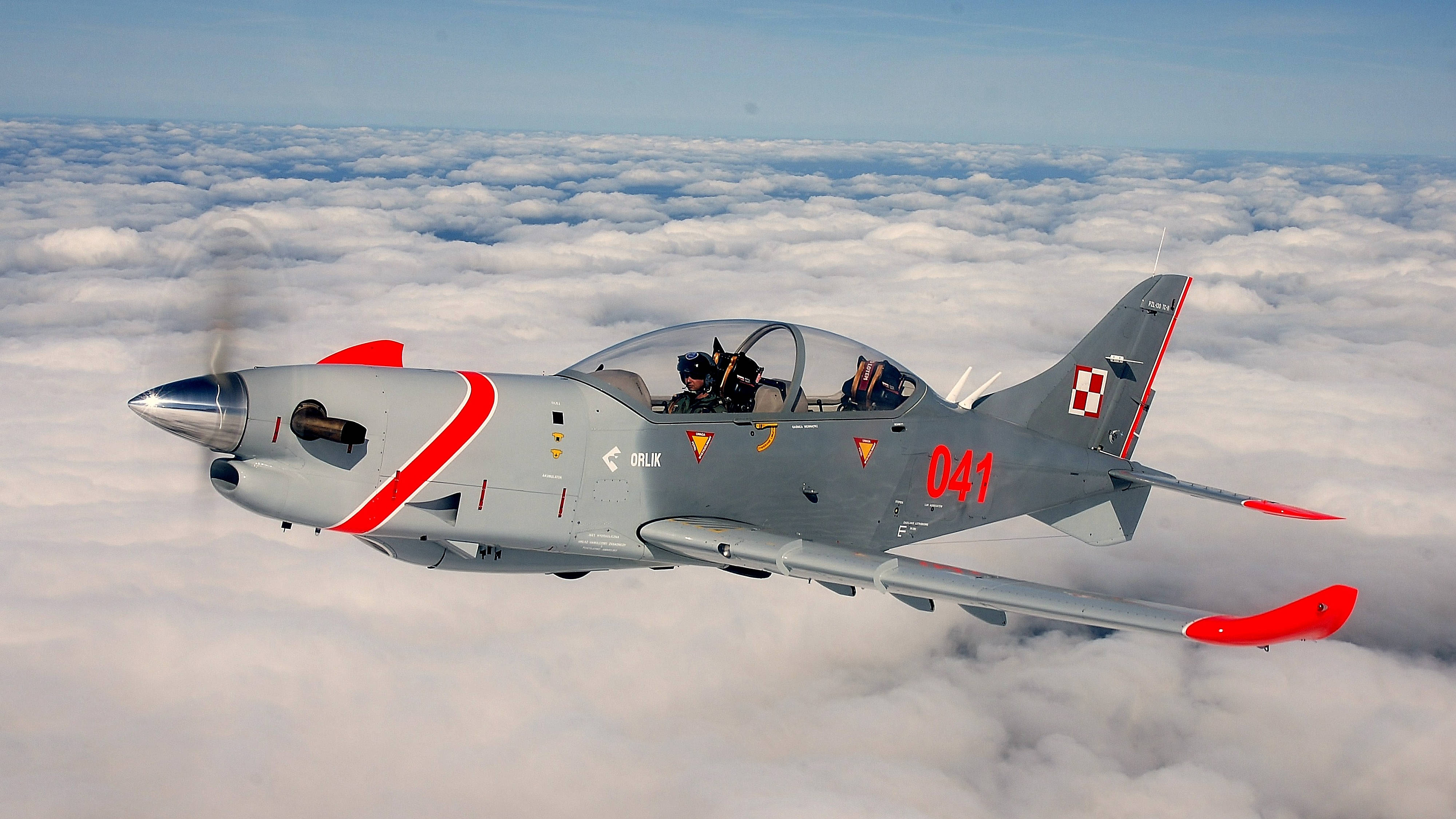 Air Force 桌布 壁紙 — 一架灰色和紅色的波蘭軍用教練機在清澈的藍天下翱翔於廣闊的蓬鬆白雲之上。