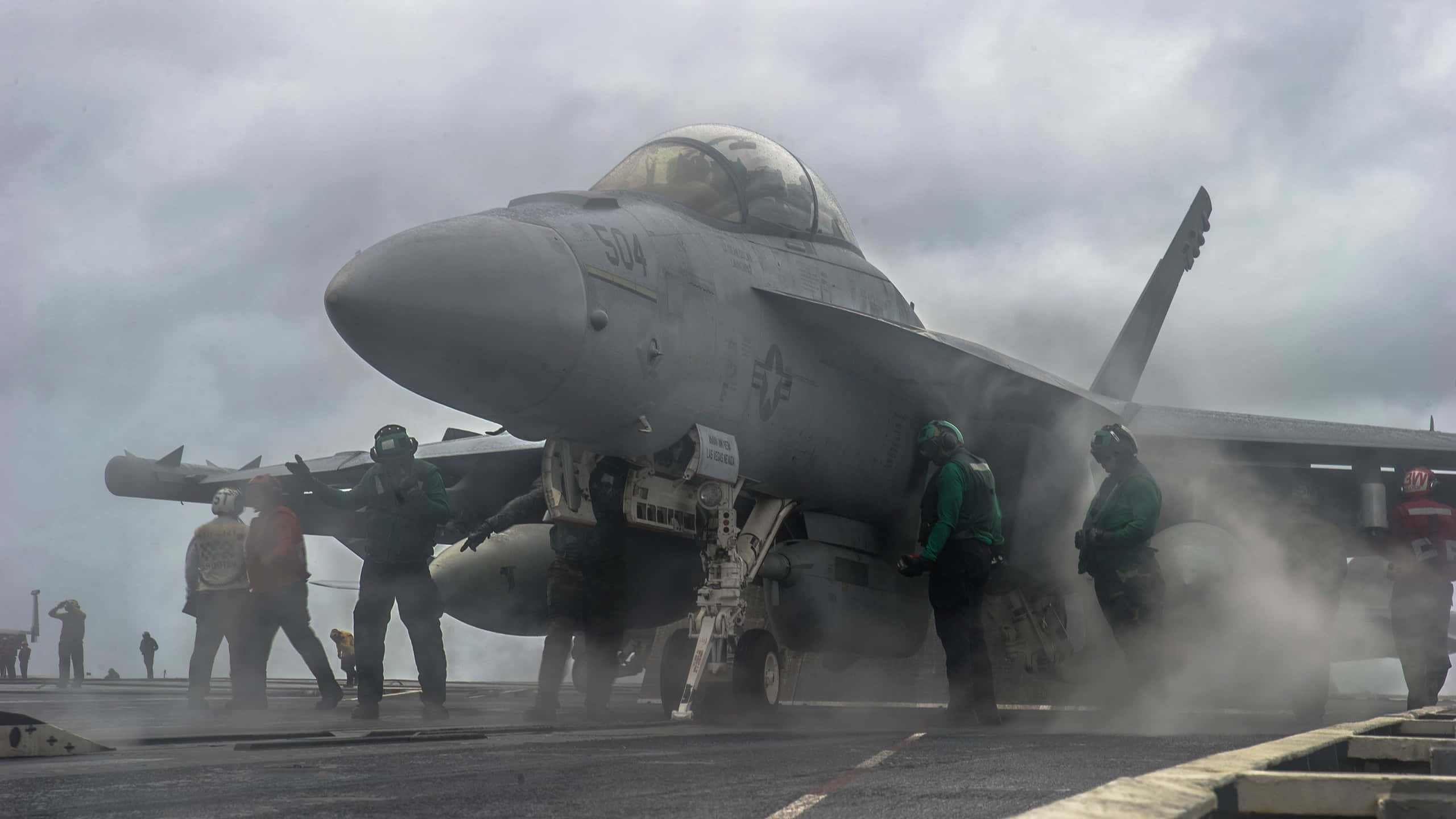 軍事動圖 桌布 壁紙 — 一架灰色戰鬥機在航空母艦甲板上，身穿綠色裝備的機組人員在陰天環境下於其周圍工作。