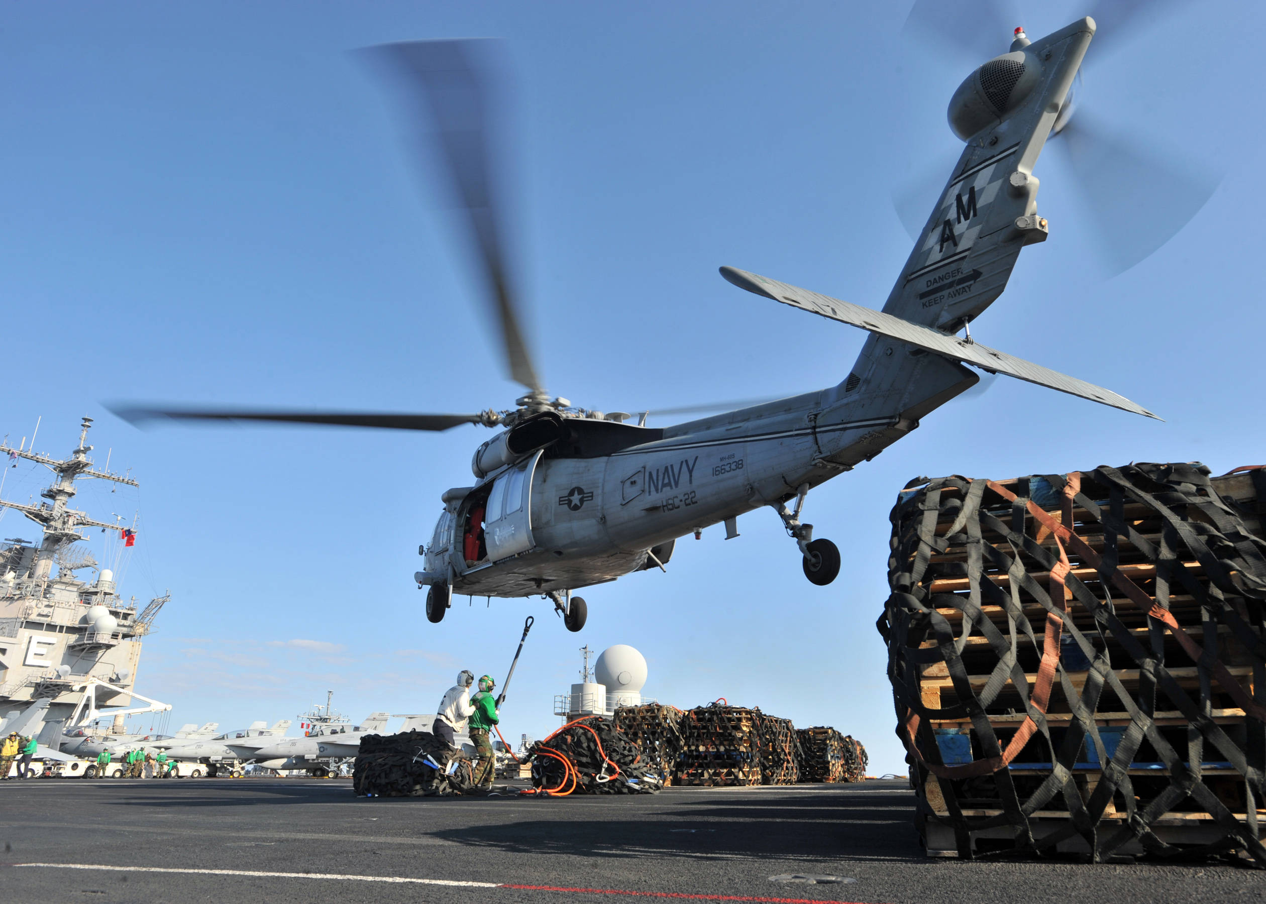 HD Military 桌布 壁紙 — 一架灰色海軍直升機在航空母艦甲板上盤旋，船員在晴朗藍天下處理貨物。