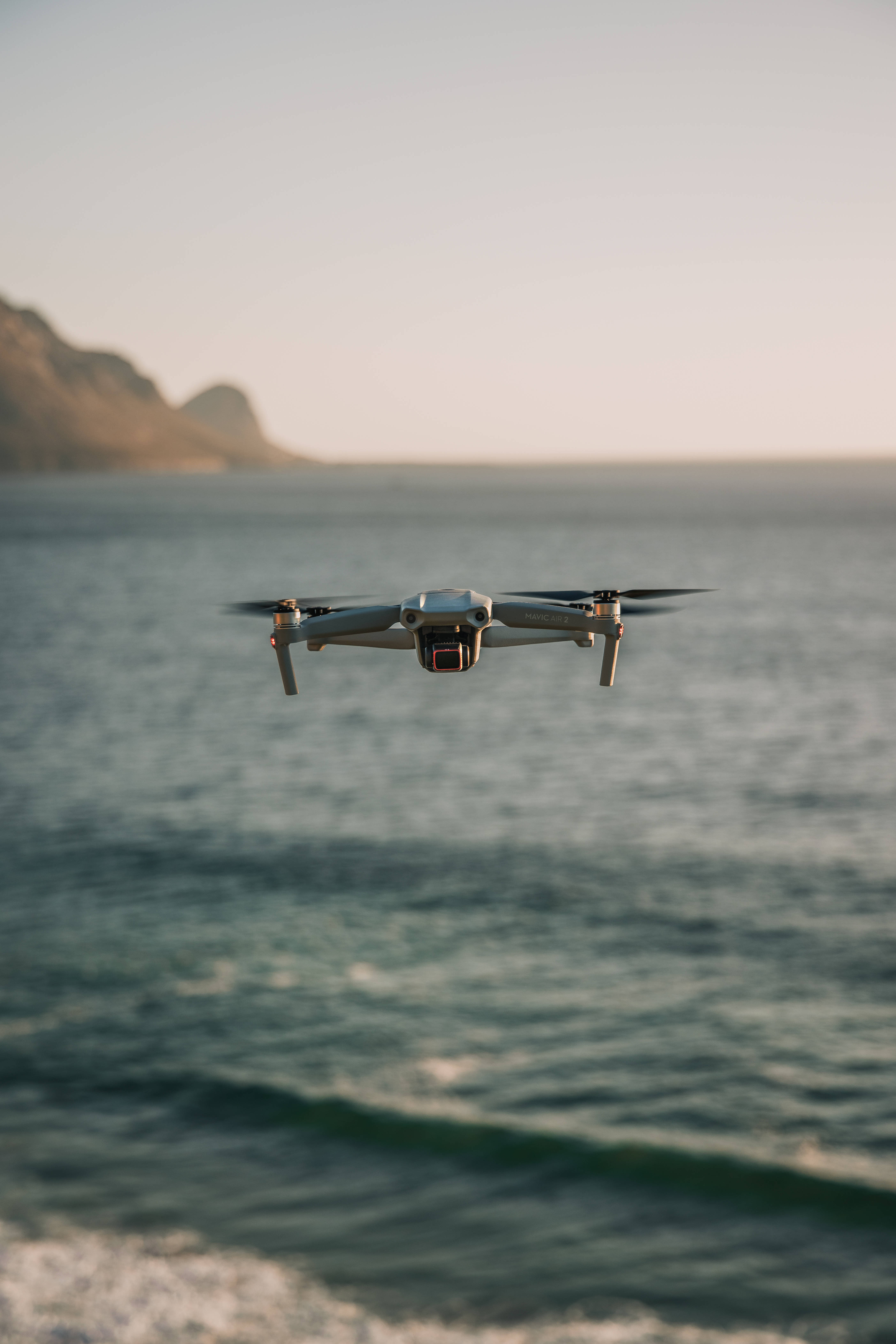 high 桌布 壁紙 — 一架灰色無人機在平靜的海洋上空盤旋，遠方懸崖襯托著柔和的金色日落天空。