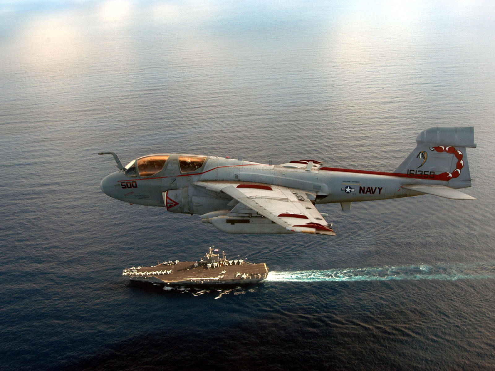 飛機 桌布 壁紙 — 一架灰色的海軍EA-6B徘徊者式飛機低飛過海洋，下方有一艘航空母艦留下白色尾跡。