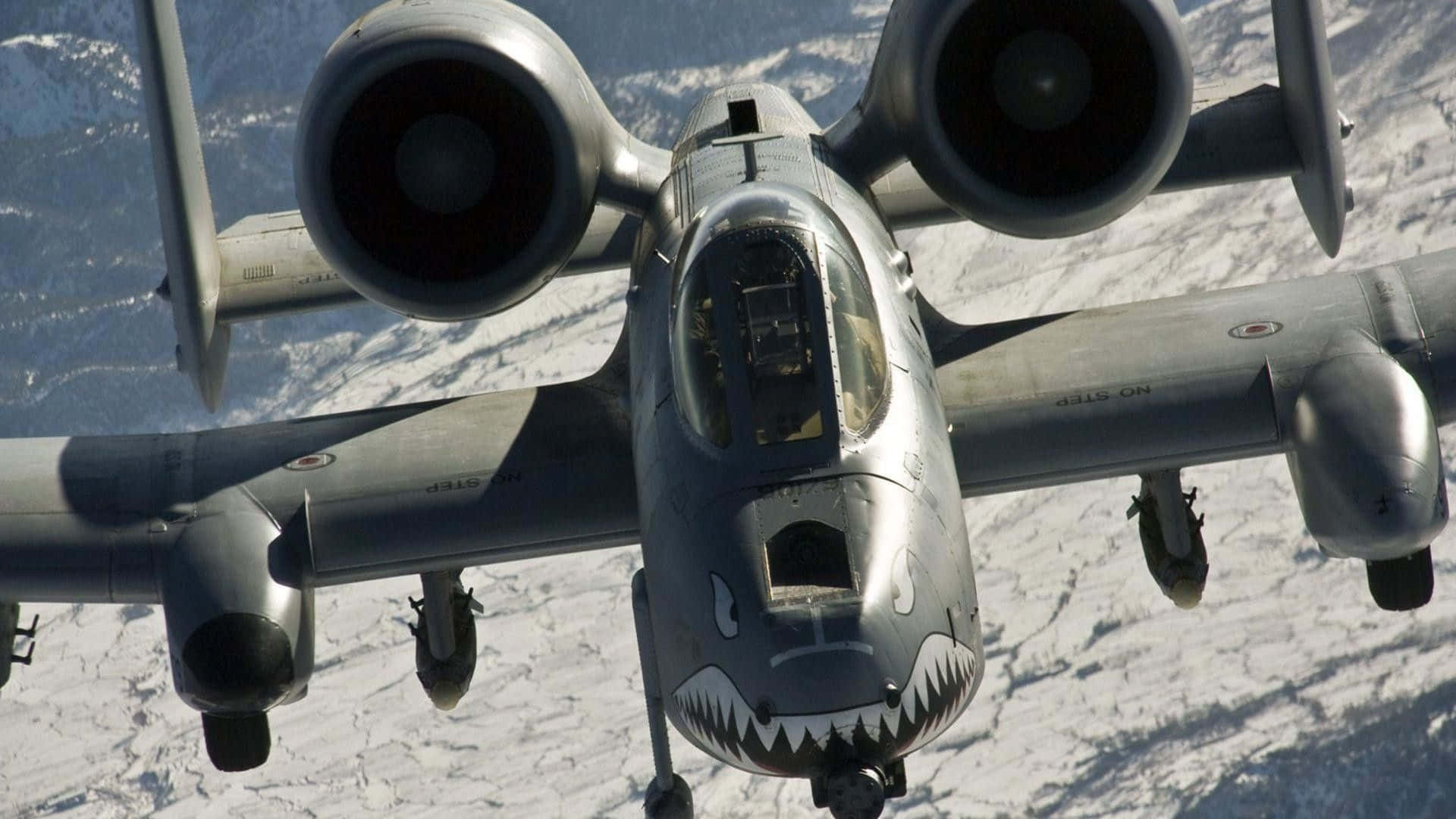 A 10 Warthog 桌布 壁紙 — 一架灰色軍用攻擊機帶有鯊魚嘴鼻藝術，在明亮的日光下翱翔於積雪覆蓋的山脈之上。