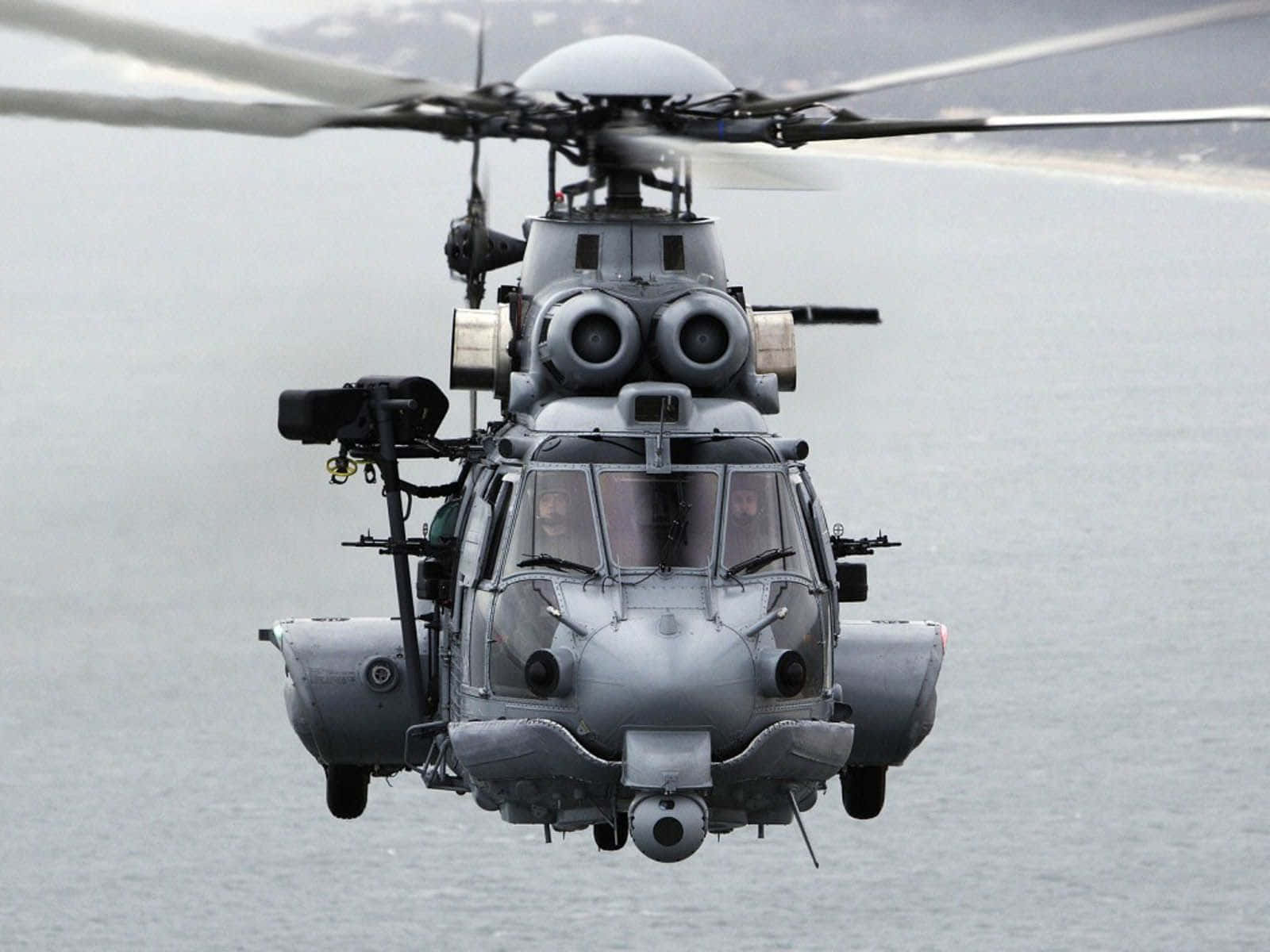 直升機 桌布 壁紙 — 一架灰色軍用直升機低空盤旋在平靜的海面上，旋翼槳葉因運動而模糊。