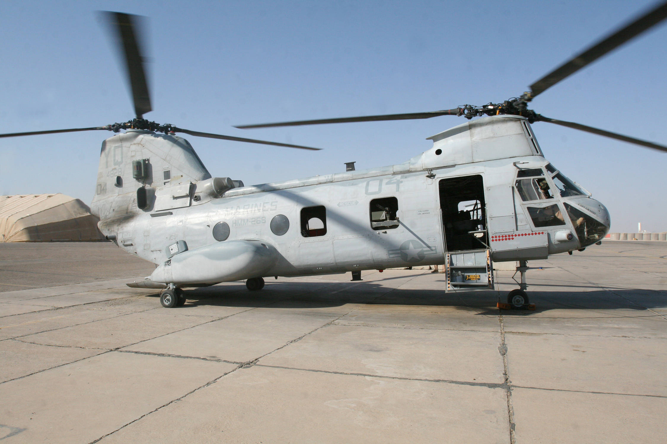 Helicopter 桌布 壁紙 — 一架灰色軍用直升機側門打開，靜止在混凝土停機坪上，天空清澈湛藍。