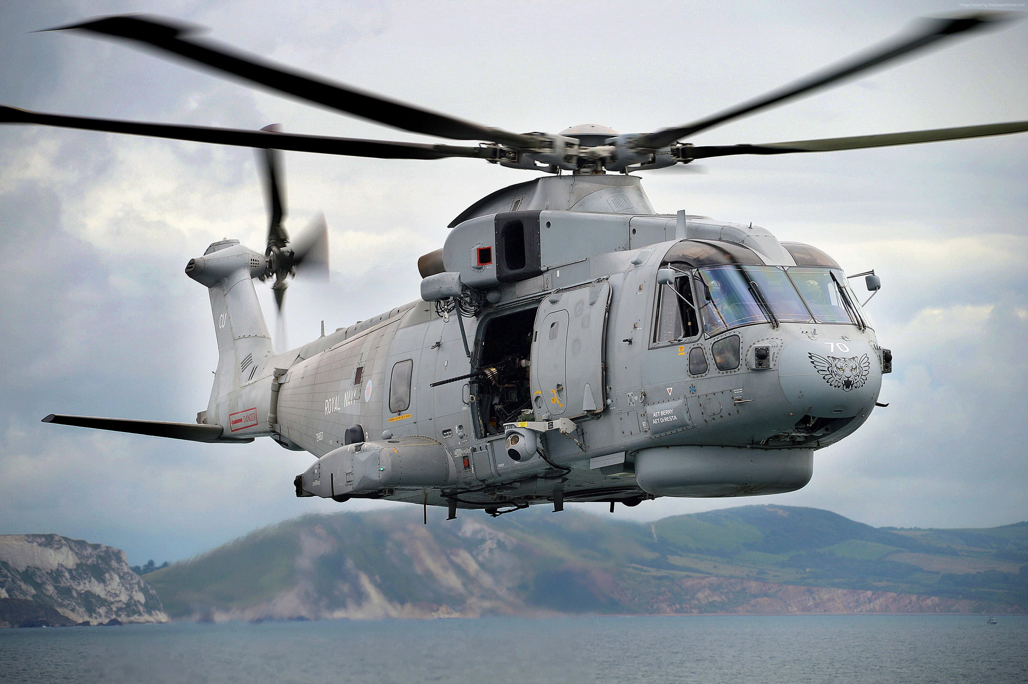 4K Helicopters 桌布 壁紙 — 一架灰色軍用直升機在陰天背景下，盤旋於沿海水域上空，背景是起伏的綠色山丘。