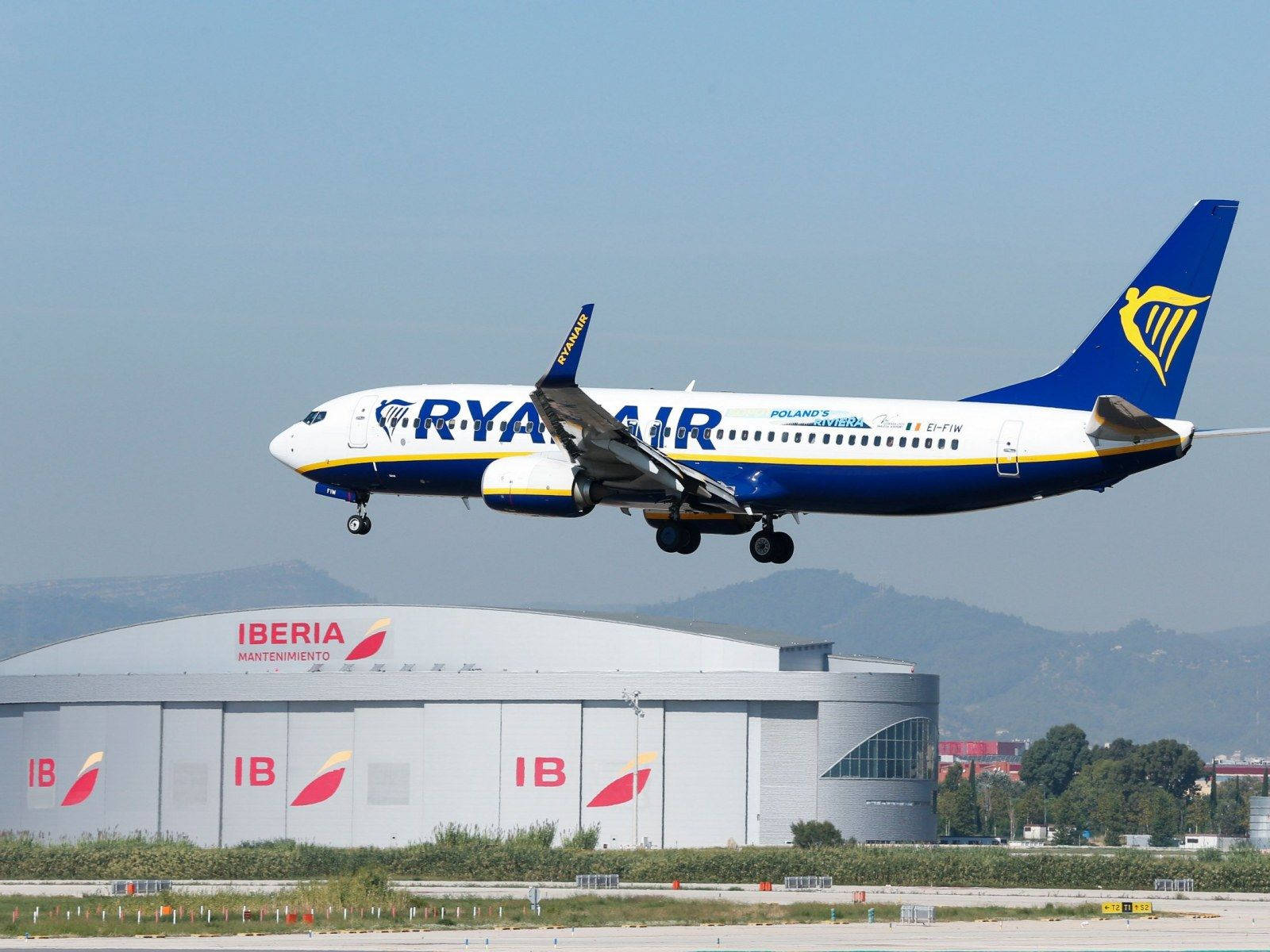 flying 桌布 壁紙 — 一架瑞安航空波音737從機場跑道起飛，背景有伊比利亞航空維修機庫和遠山。