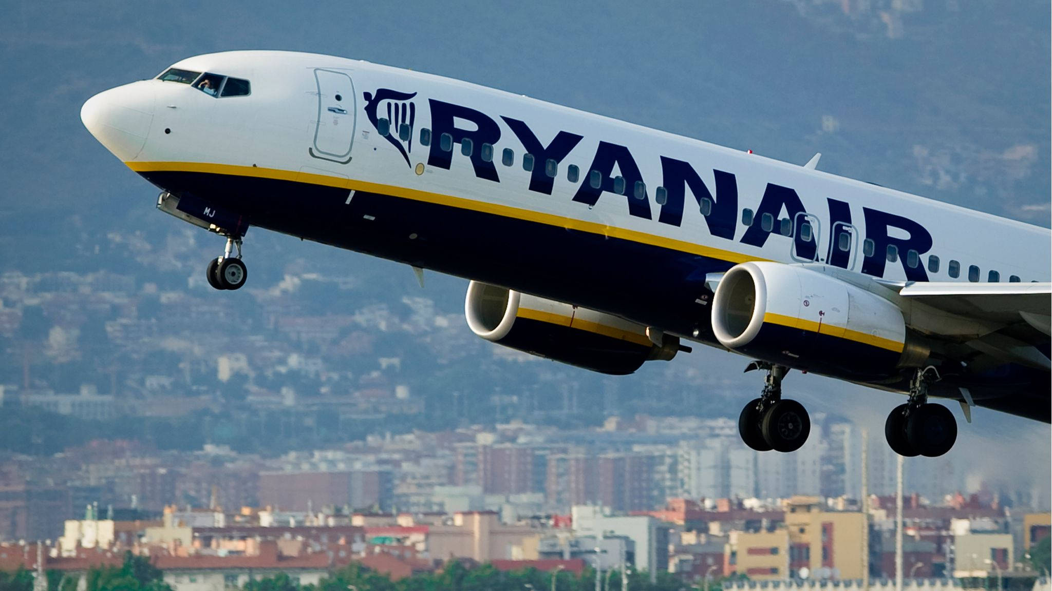 飛機 桌布 壁紙 — 一架瑞安航空的飛機正在飛行中，起落架已放下，在晴朗的天空下翱翔於廣闊的城市景觀之上。