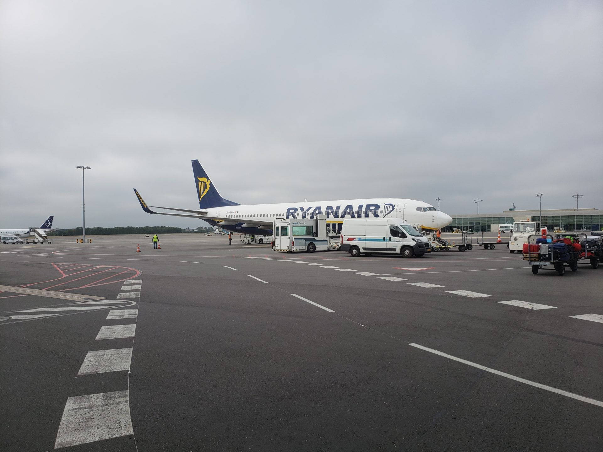 飛機 桌布 壁紙 — 一架瑞安航空飛機停泊在機場閘口，天空多雲，附近有地勤人員和服務車輛。