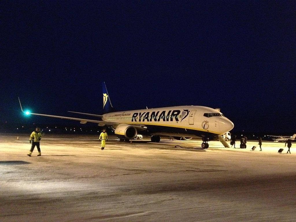 飛機 桌布 壁紙 — 一架瑞安航空飛機在夜間停在積雪的停機坪上，身穿黃色外套的地勤人員在人工燈光下在附近行走。