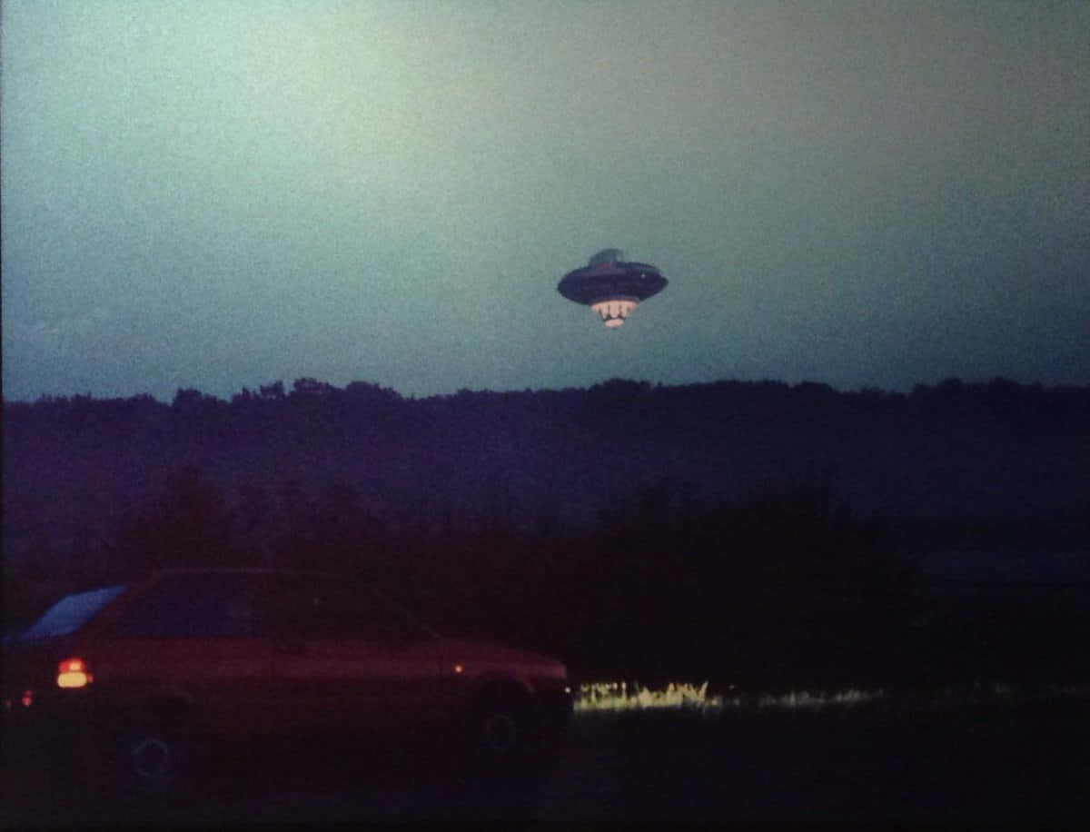 外星人 桌布 壁紙 — 一架發光的UFO懸浮在黑暗的森林上空，一輛汽車的尾燈在黃昏的路邊閃爍。