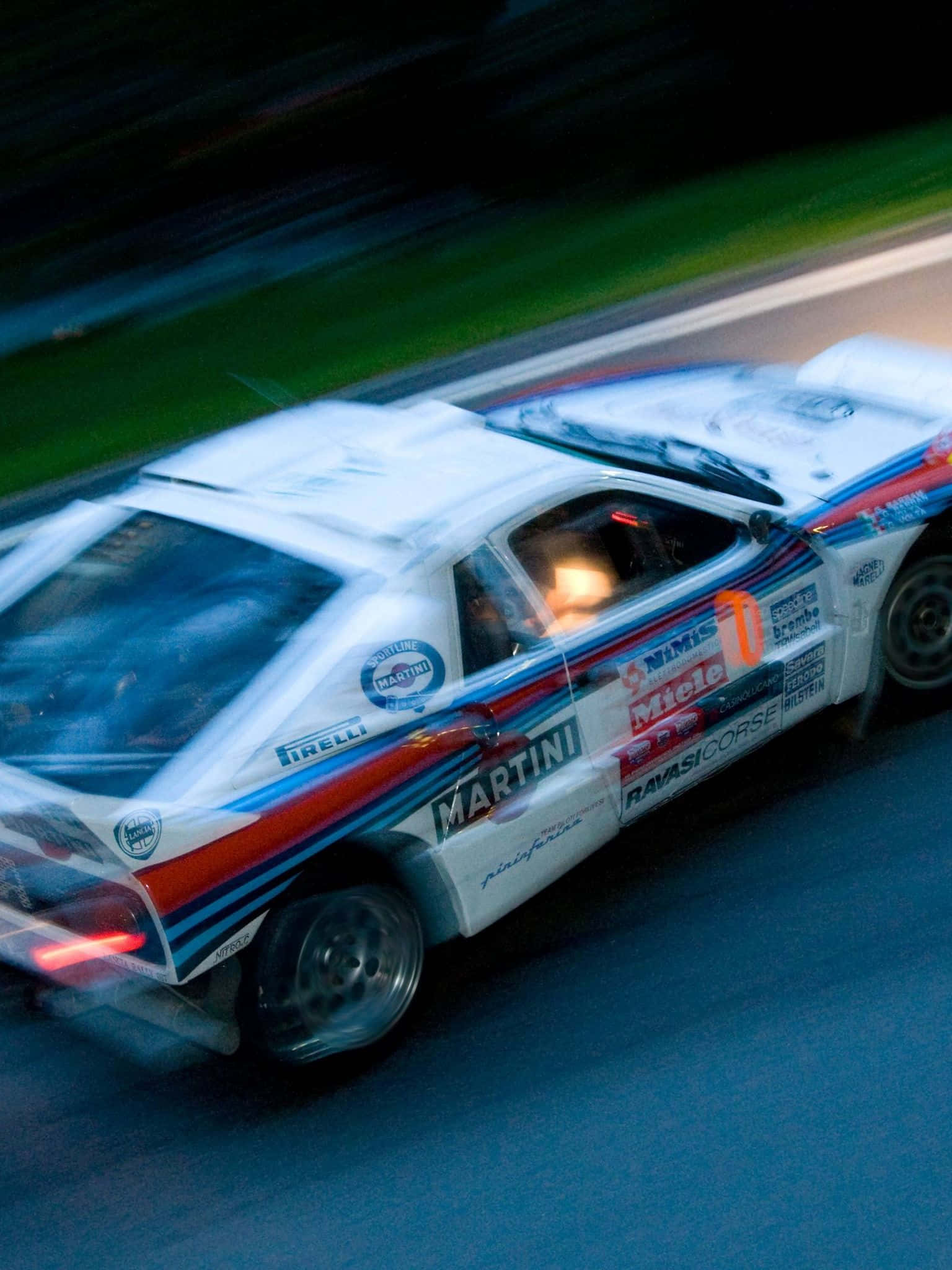 Lancia 桌布 壁紙 — 一架白色、藍色和紅色的馬丁尼賽車在夜間賽道上模糊駛過，帶有發光的車頭燈和動態條紋。