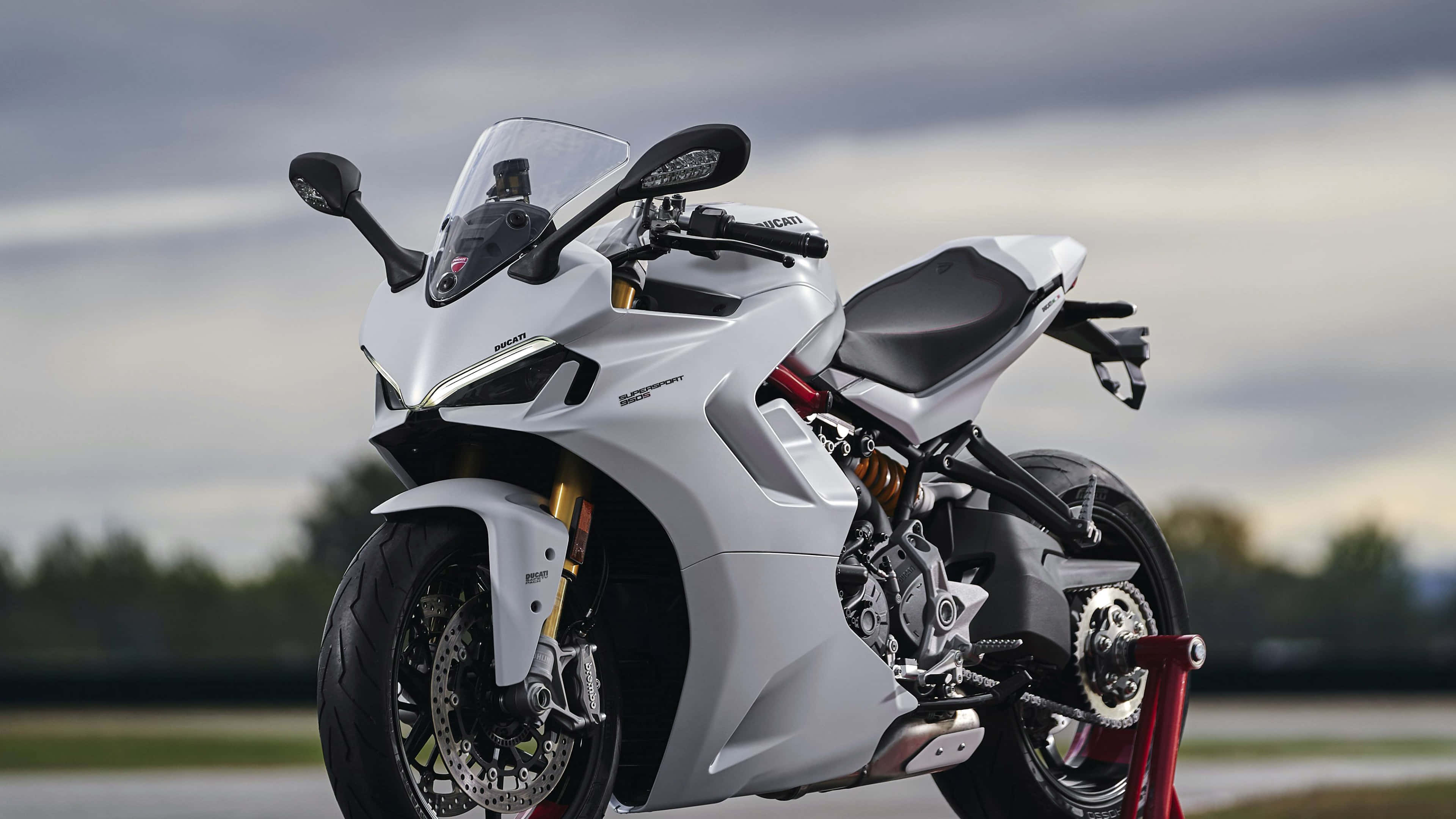 桌布 壁紙 — 一架白色Ducati機車，配金色前叉和黑色座椅，在紅色支架上，背景是陰天。