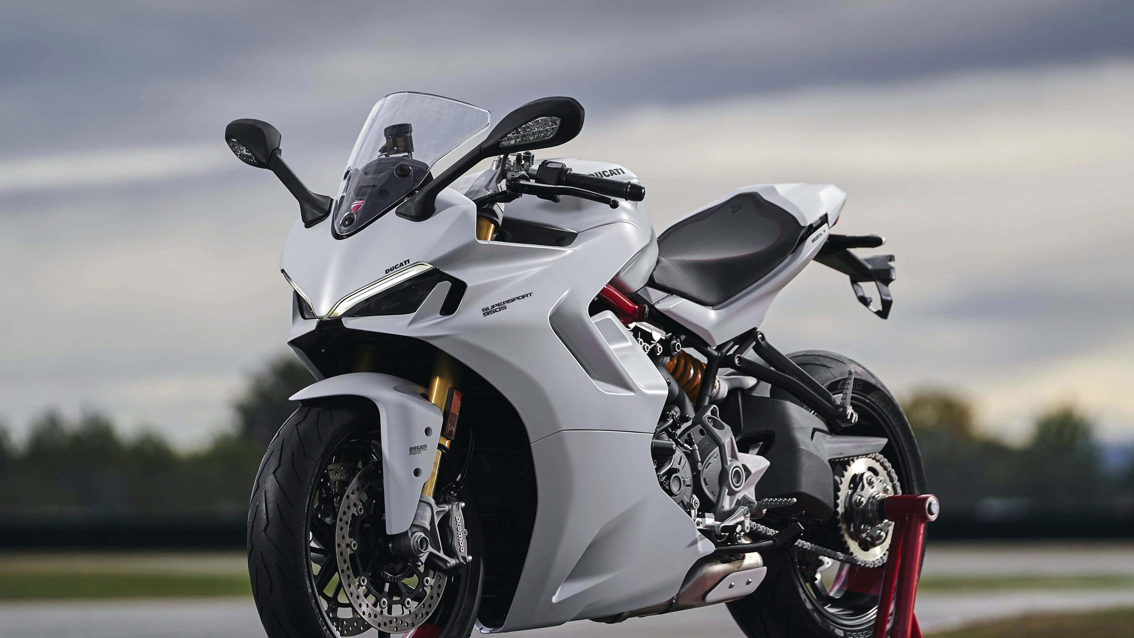 4K Bikes 桌布 壁紙 — 一架白色Ducati運動機車優雅地停放在紅色支架上，背景是陰天和模糊的樹木。